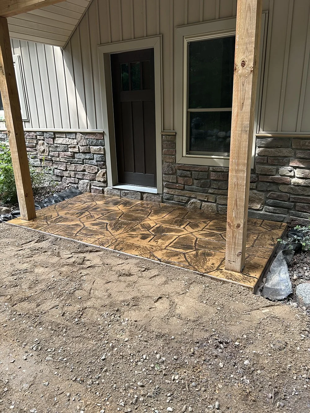 Newly built front porch with stamped concrete flooring, wooden posts, next to a house with brick and metal siding, and a closed dark front door.