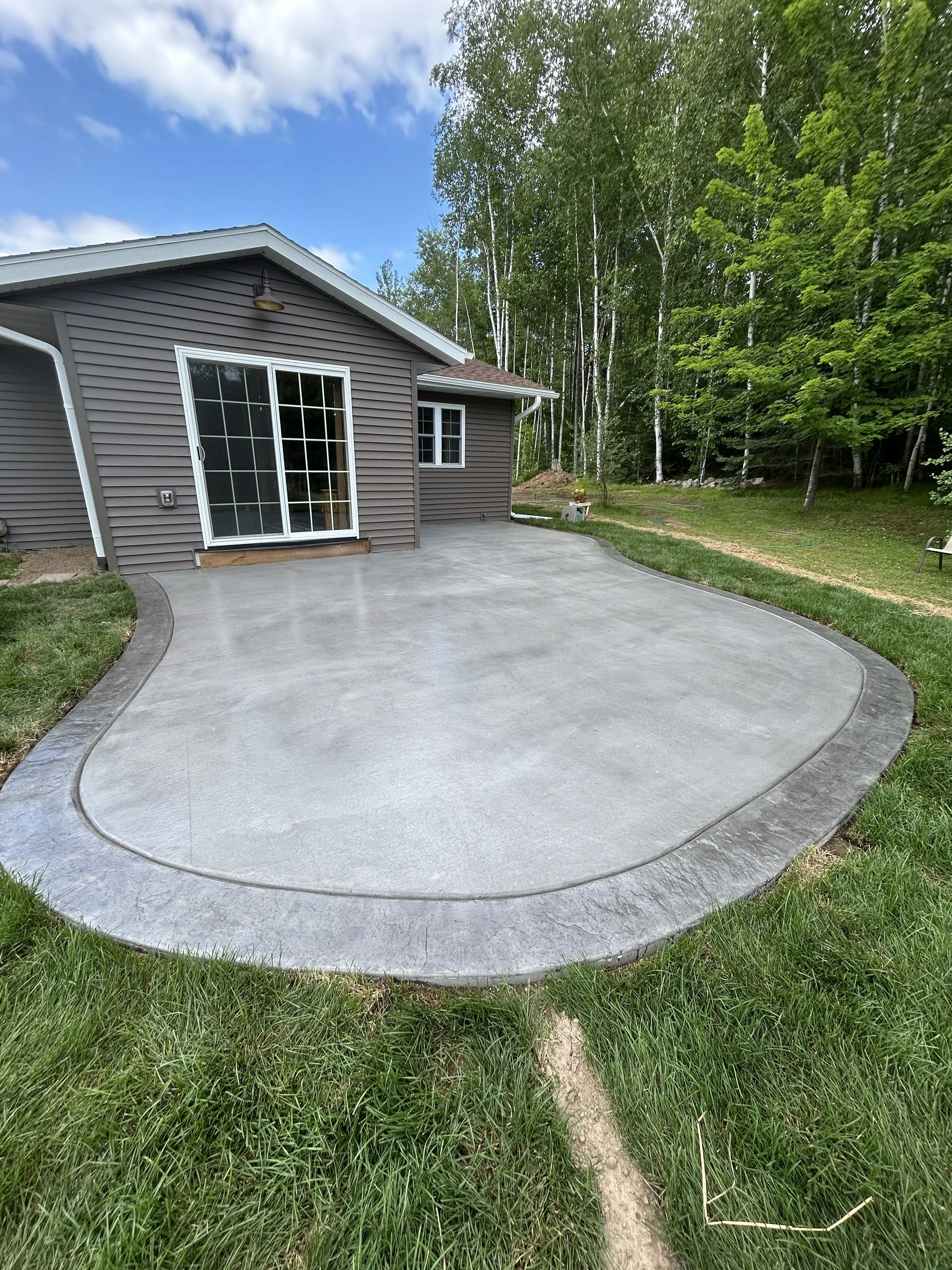 Newly poured concrete patio with a curved edge in the backyard of a house, surrounded by green grass and trees.
