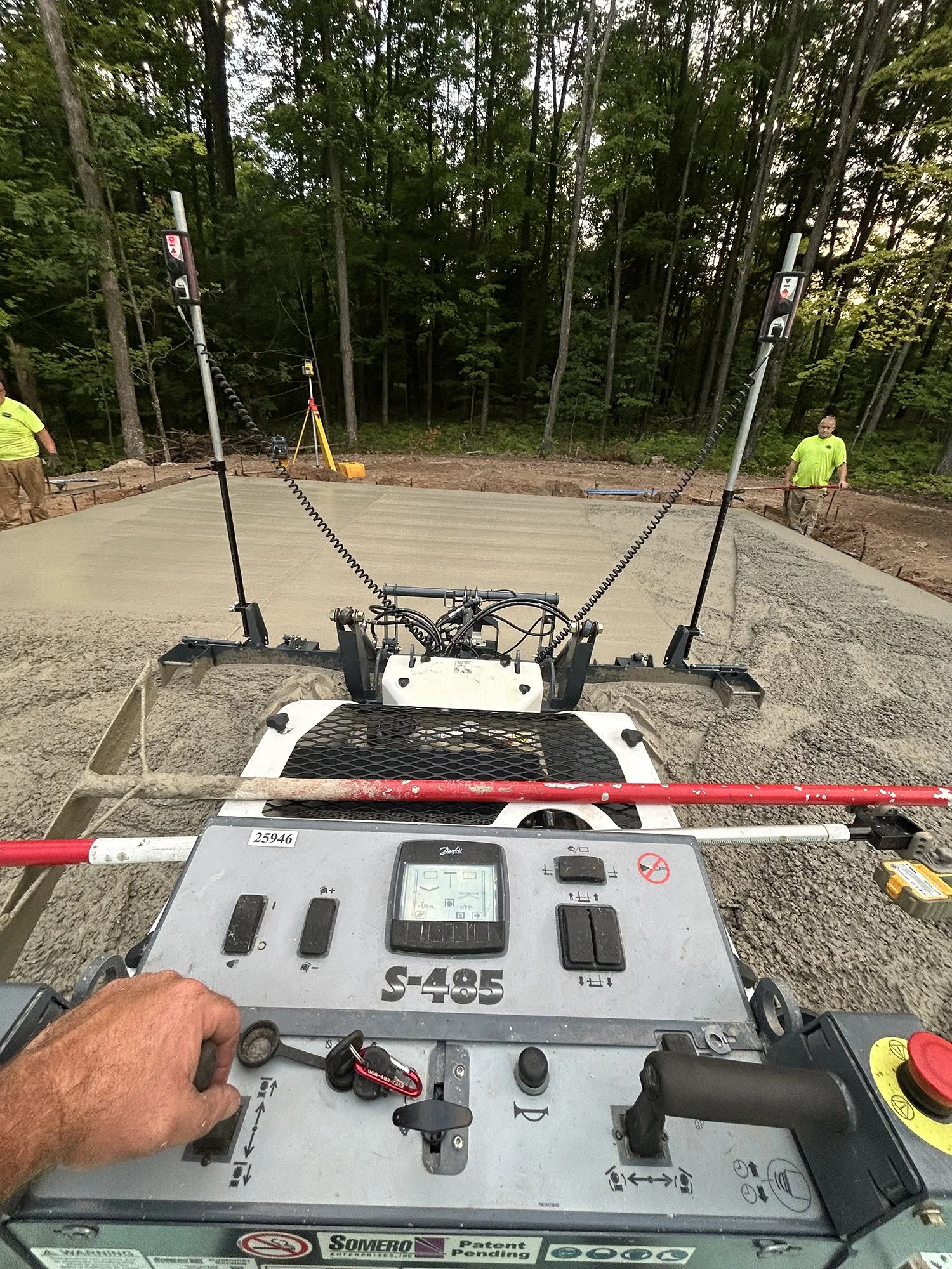 Operator using a concrete paving machine to lay a concrete slab in a forested area. Two workers in neon green shirts stand nearby supervising the process.
