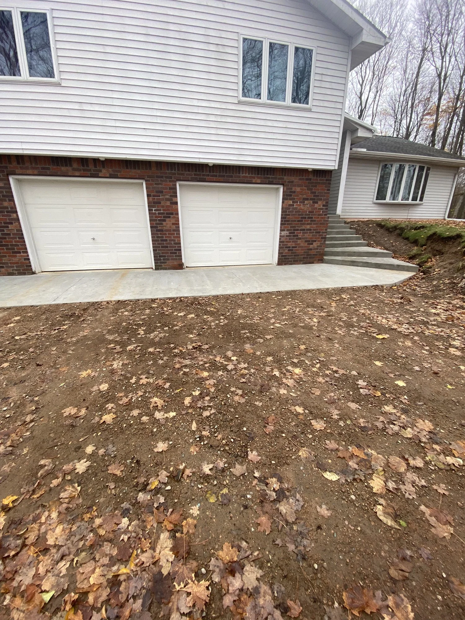 Front view of a house with a double garage, stairs on the right side, and leaf-covered dirt yard in the foreground.