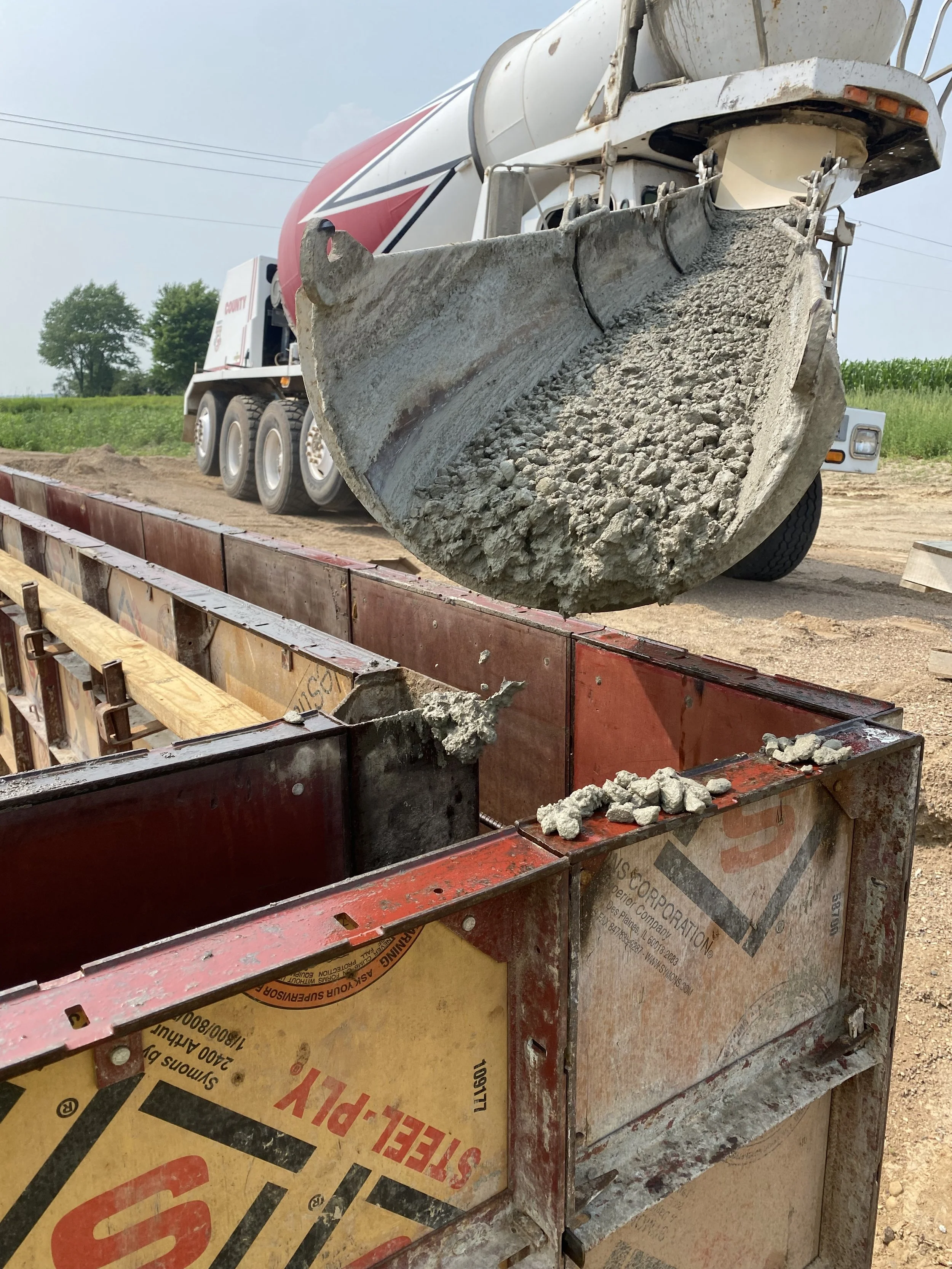 A cement mixer truck dispensing concrete into a red metal formwork on a construction site with dirt ground and green trees in the background.