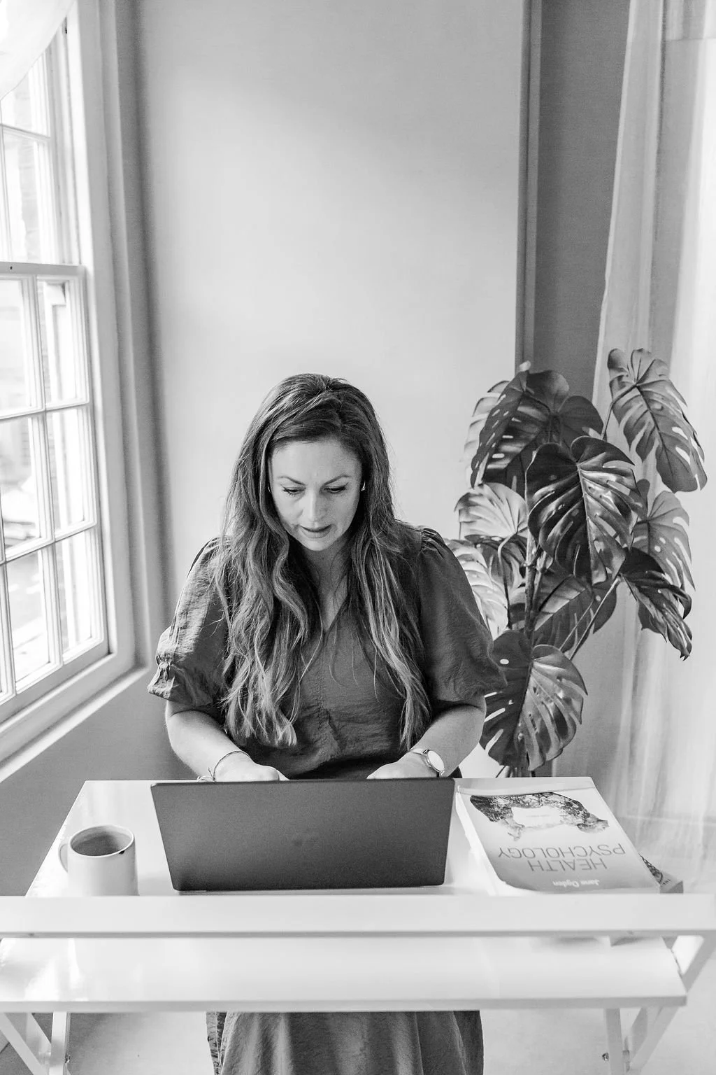 A woman sitting at a desk using a laptop, with a coffee mug and a book titled 'Healthy Dishology' on the desk, next to a large houseplant, by a window.