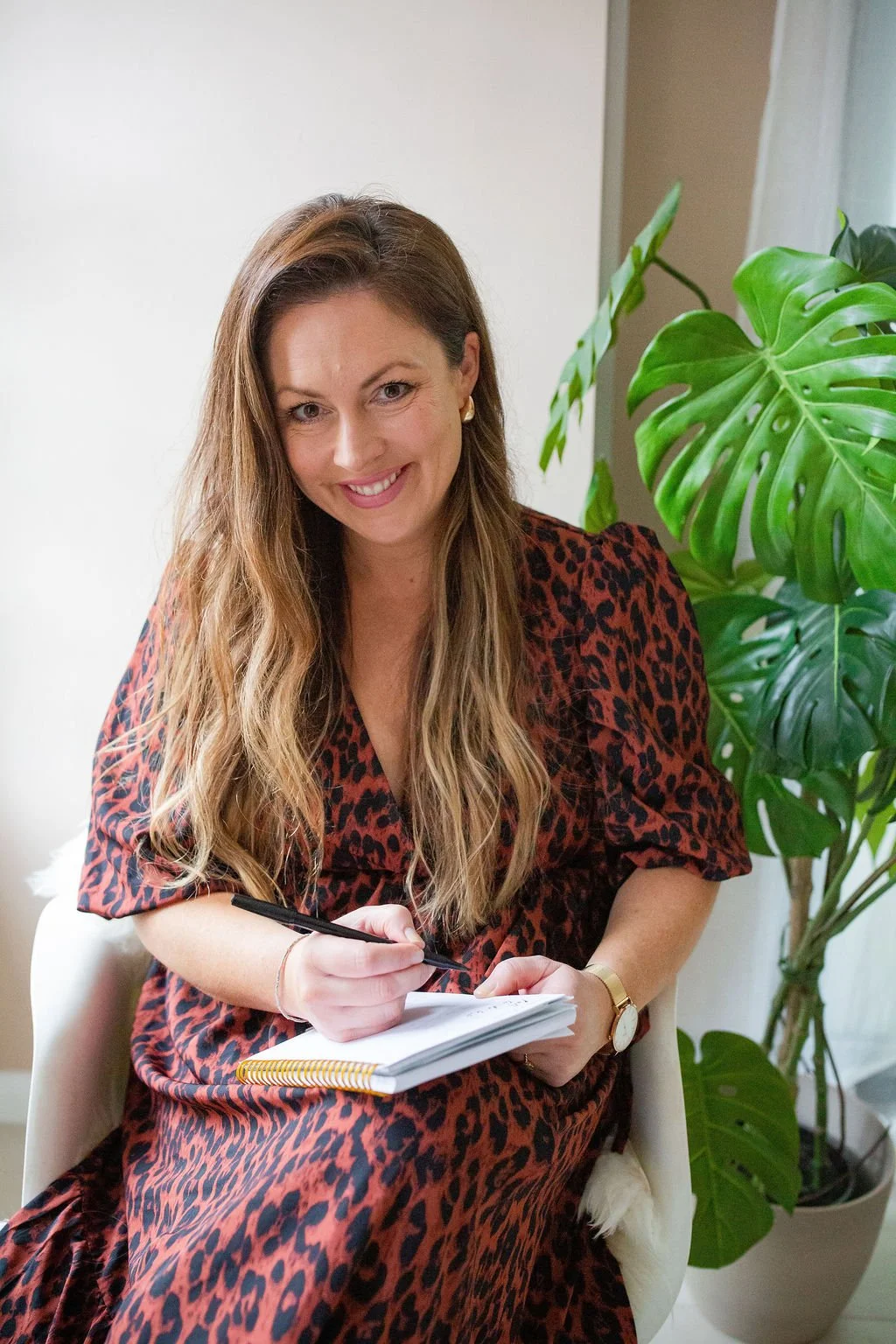 A woman with long wavy hair wearing a red leopard print dress, sitting on a chair with a notebook and pen, smiling, next to a large green potted plant.