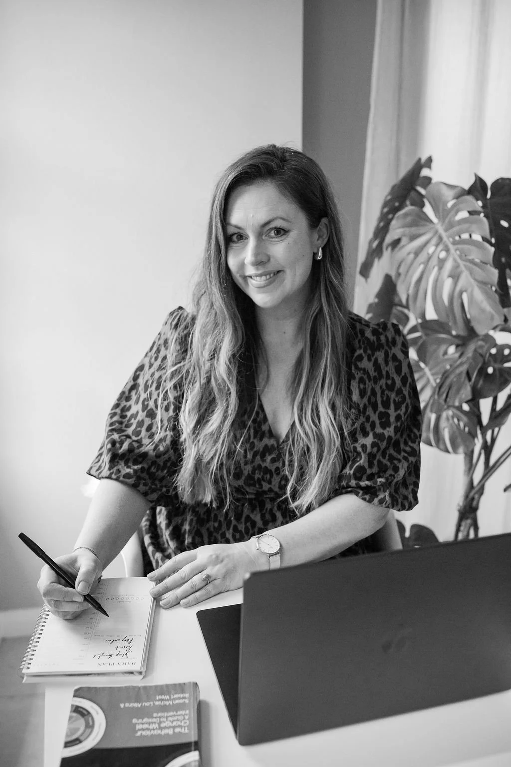 A woman with long wavy hair sitting at a desk in a meeting or working, smiling, with a notebook and pen in hand. She is wearing a leopard print top and a watch, with a laptop and a plant visible on her desk.