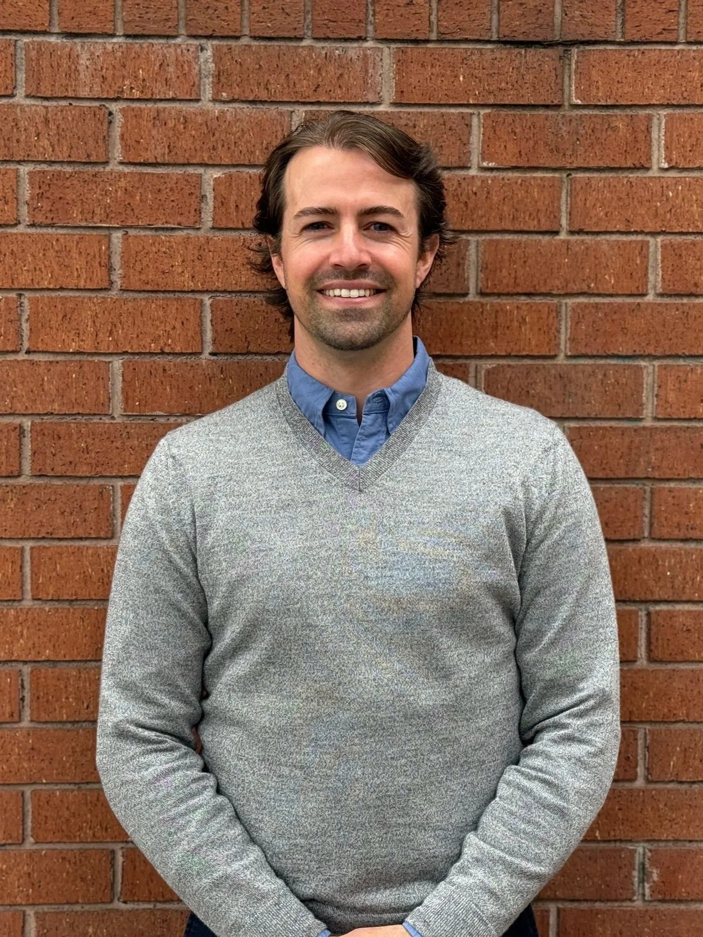 An image of Eddie Vacarro, smiling man with brown hair, wearing a blue checked shirt, standing outdoors in front of green trees.