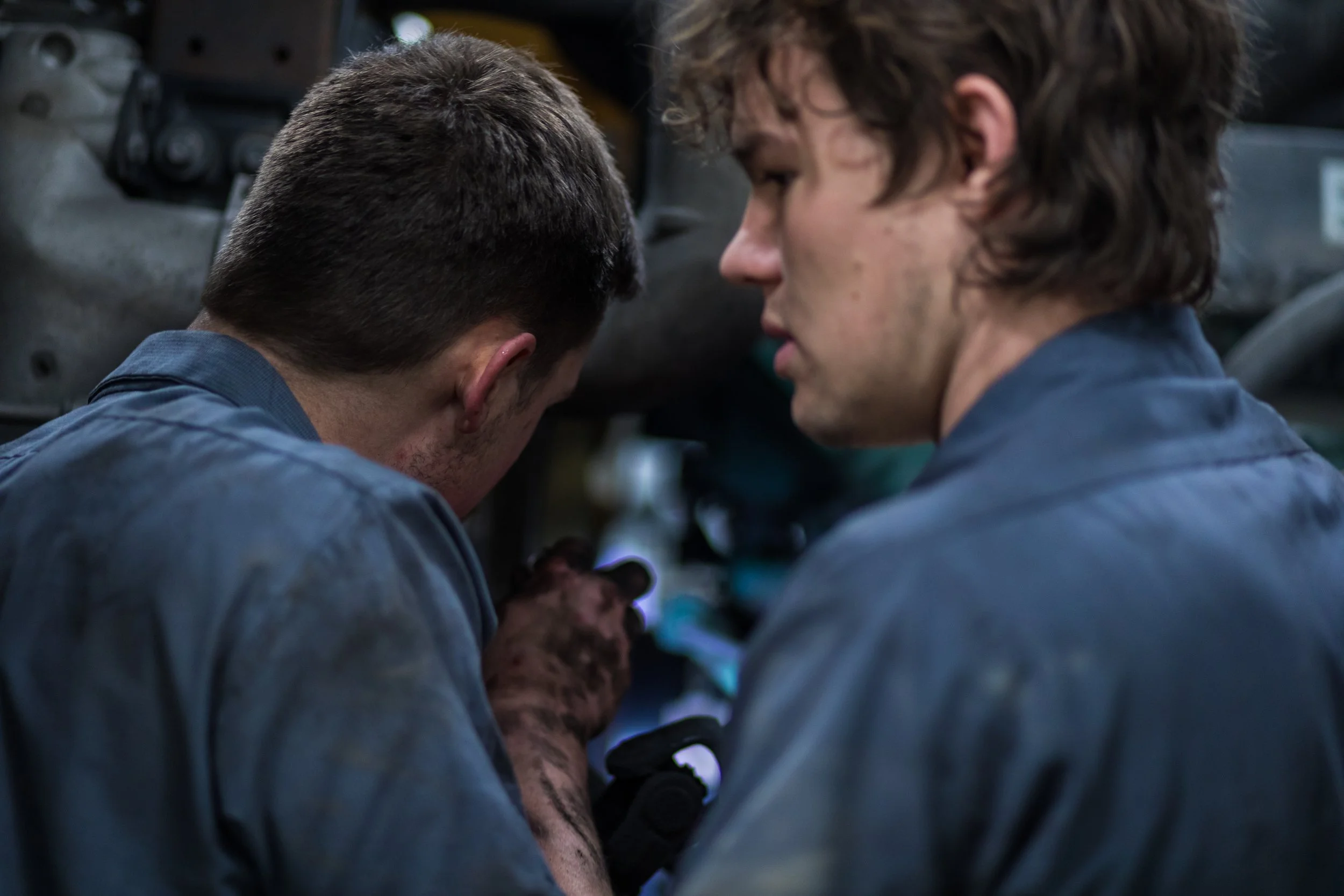 Two men in gray work uniforms in a close-up, focused on something small they are holding or working on, with a blurred mechanical background.