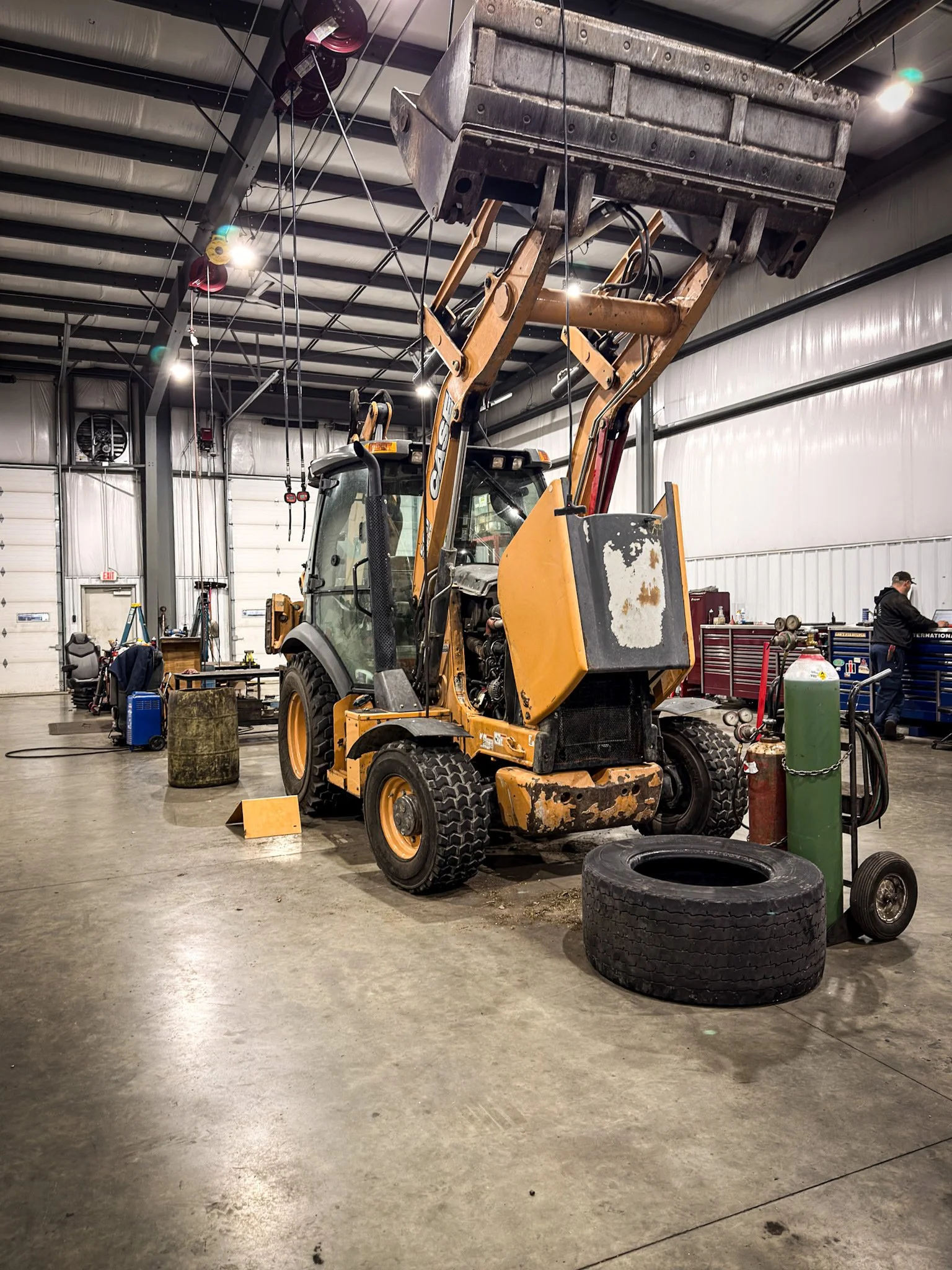 Inside a workshop with a yellow backhoe loader, tires, and various tools and equipment.