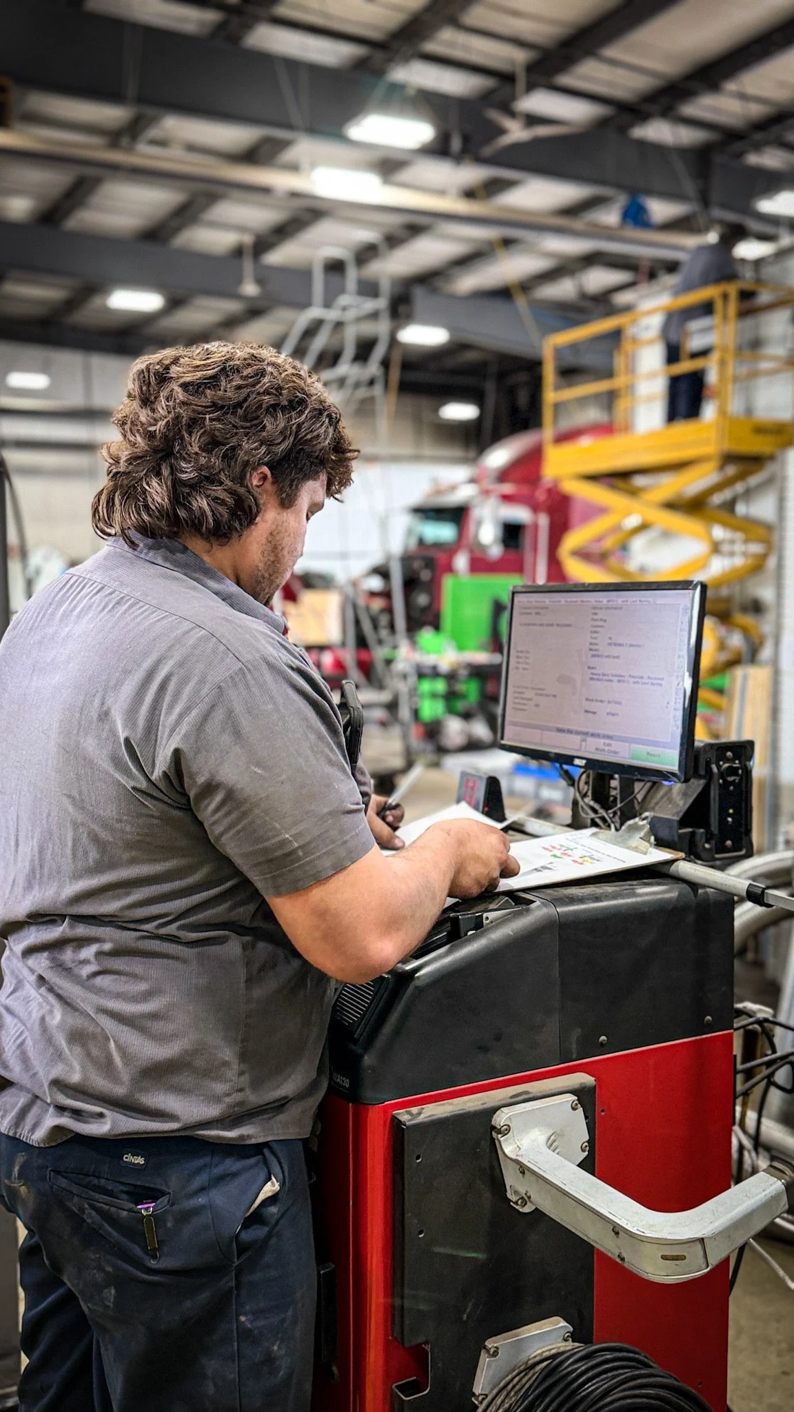 A man standing at a workstation inside an industrial workshop, working on a computer with a large monitor, surrounded by machinery, tools, and equipment, with a red truck and yellow maintenance lift in the background.