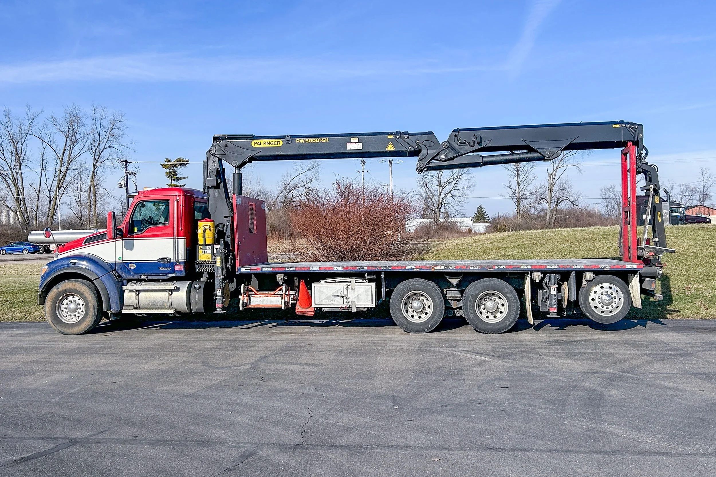 A flatbed tow truck with a mounted crane parked on an asphalt lot with grass and trees in the background.
