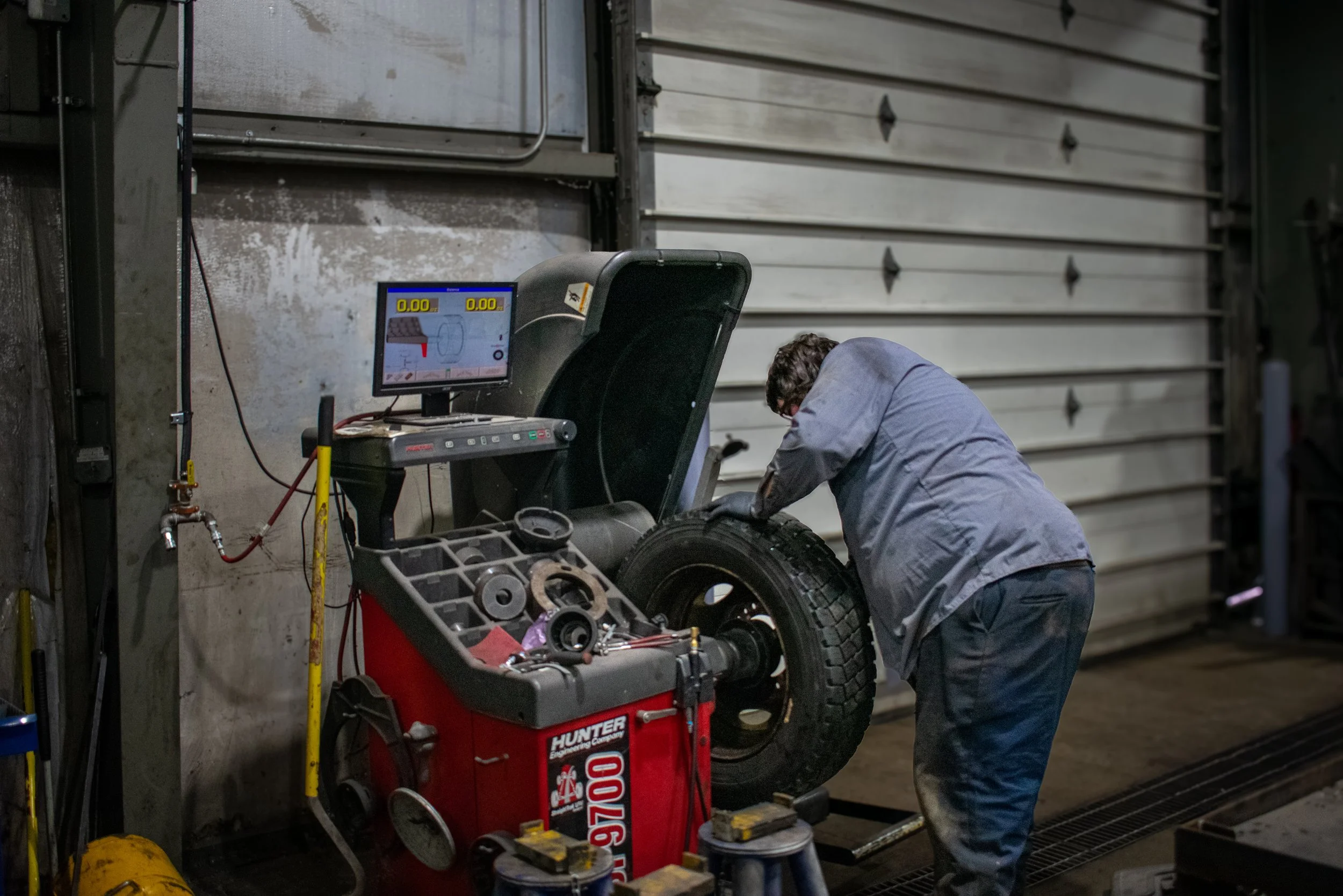 A mechanic inspects the tire of a vehicle inside an industrial garage. The garage has a closed metal roll-up door, and various tools and a computer-based wheel balancing machine are visible.