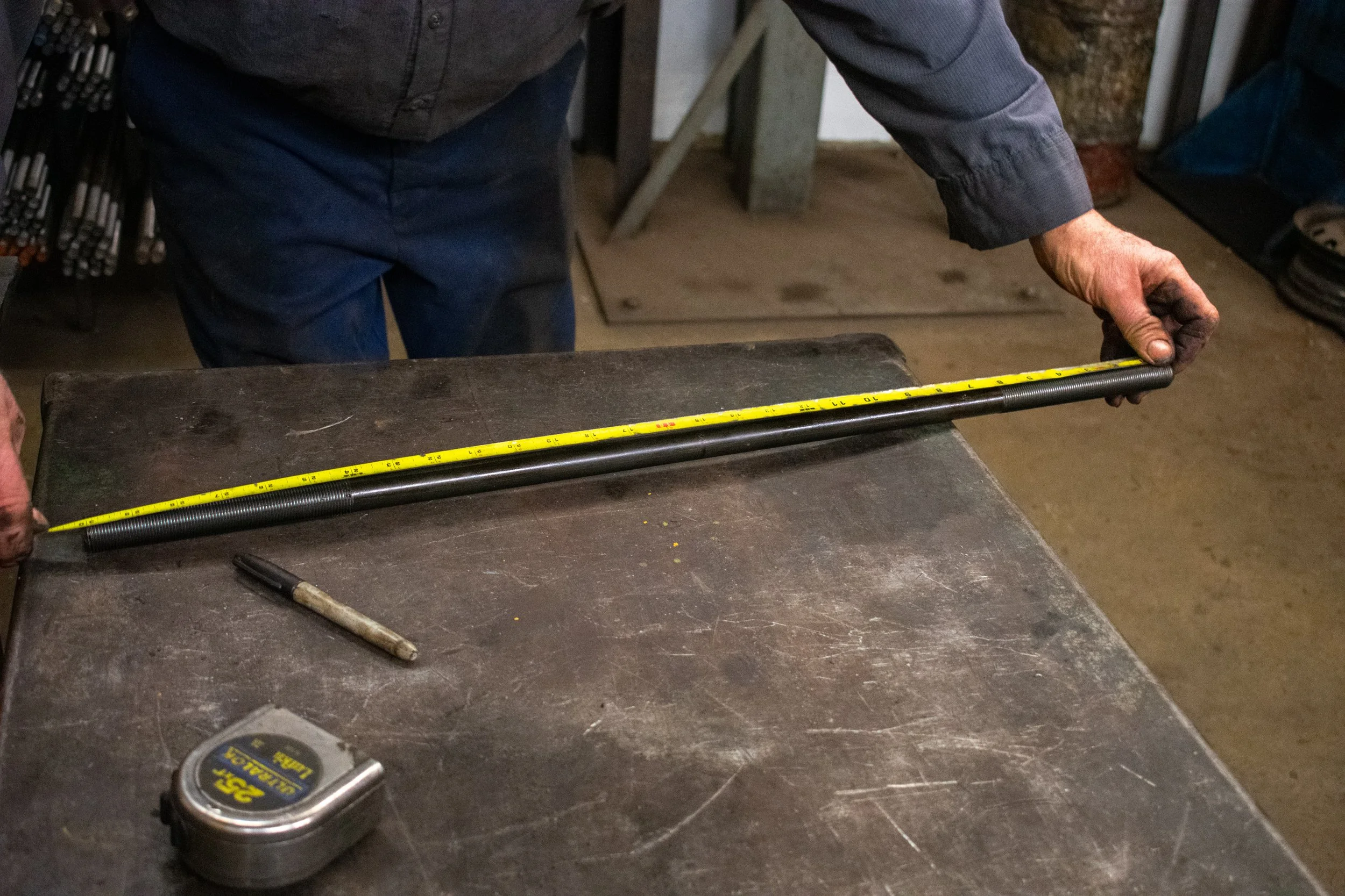 A person is measuring a long metal rod with a yellow tape measure on a workbench, with a marker and a tape measure case nearby.