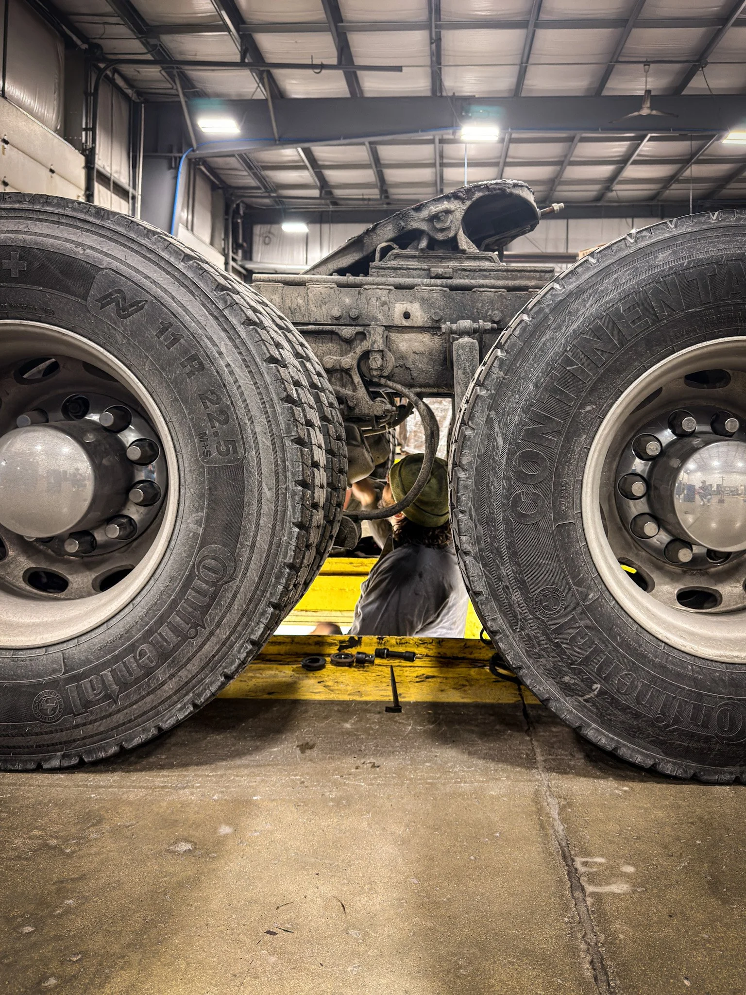 A person working underneath a large truck in a garage, with two oversized tires visible in the foreground.