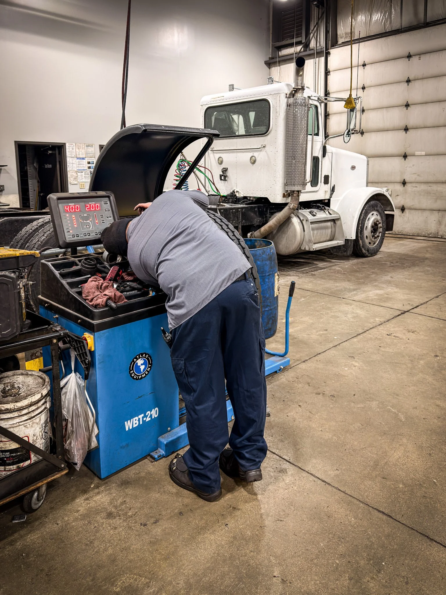 A mechanic working on a truck's tire in a garage or workshop, with a large white truck in the background and the worker bent over a tire balancing machine.