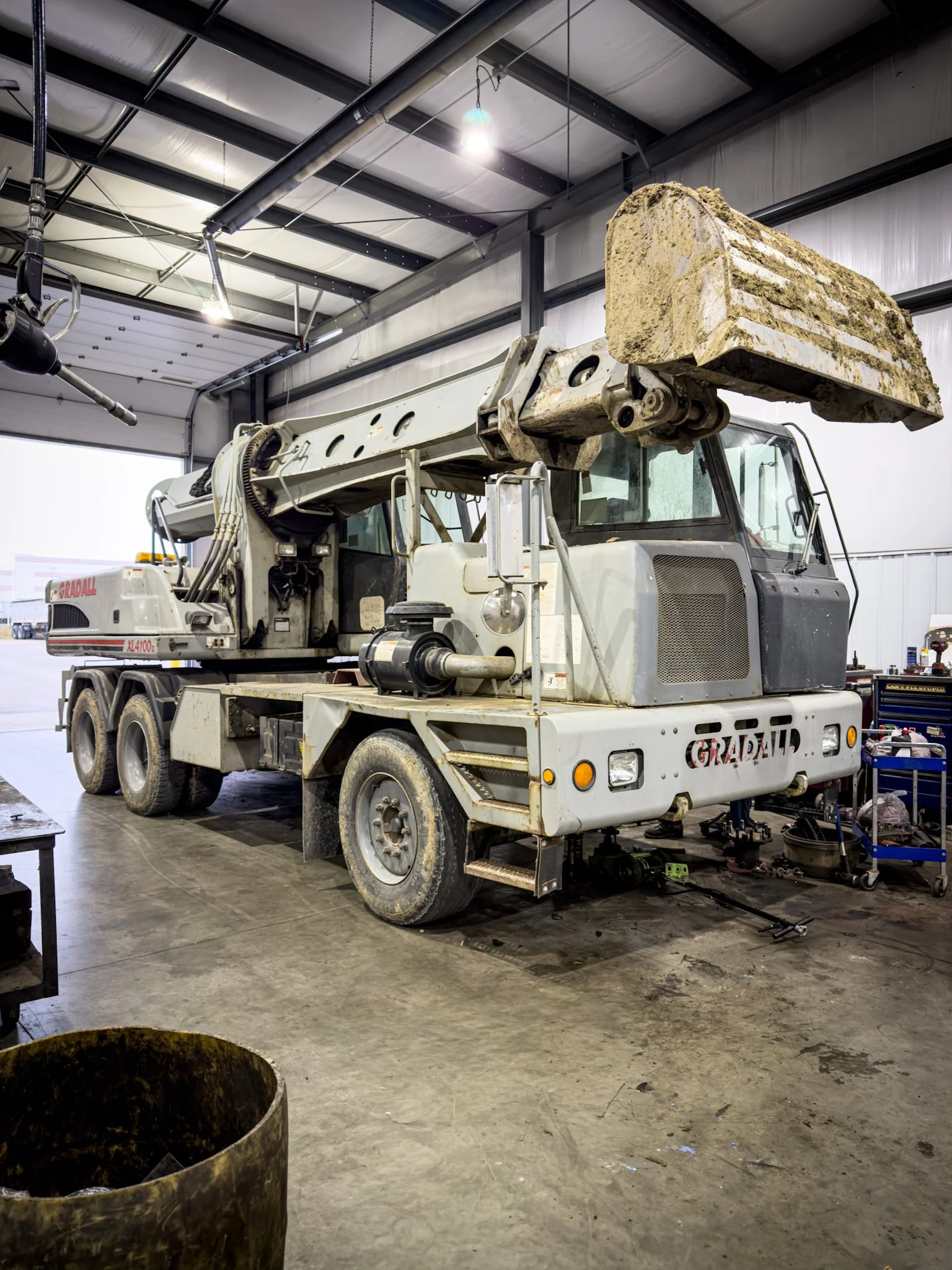 A large white industrial crane inside a warehouse, with a raised arm and a bucket attachment on top, parked on a concrete floor.