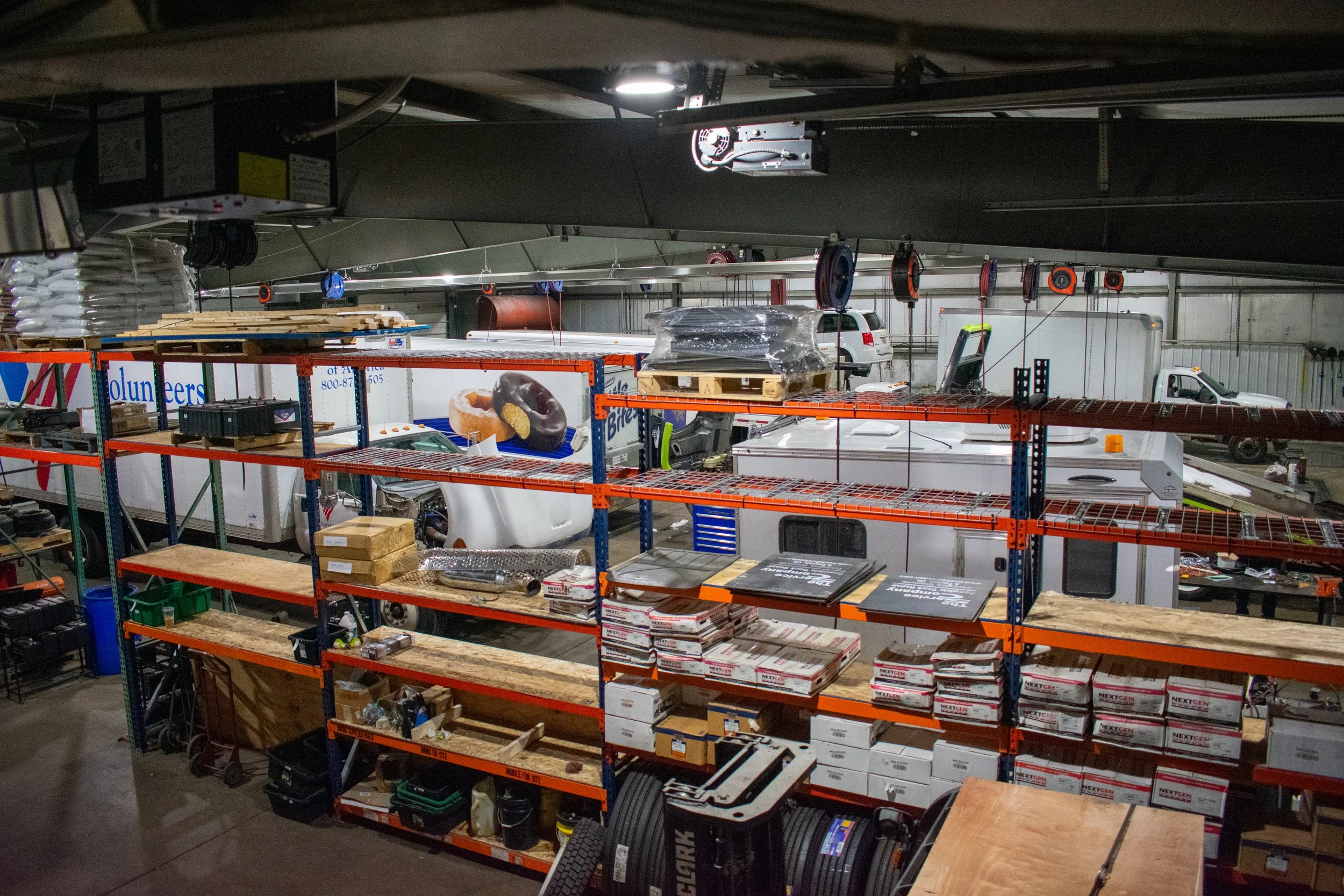 Interior of a warehouse or workshop with shelving storing boxes and equipment, multiple pickup trucks, and various tools and materials.
