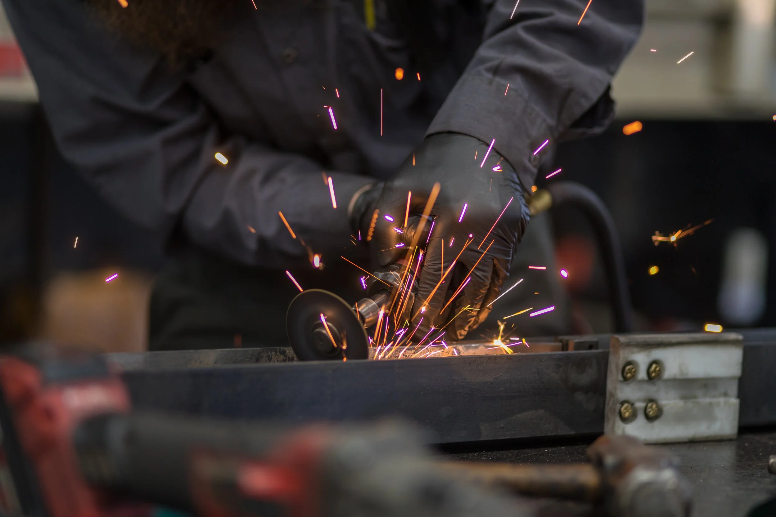 A person in black gloves and jacket is using a grinder tool, producing sparks, to cut or grind a metal piece on a workbench.