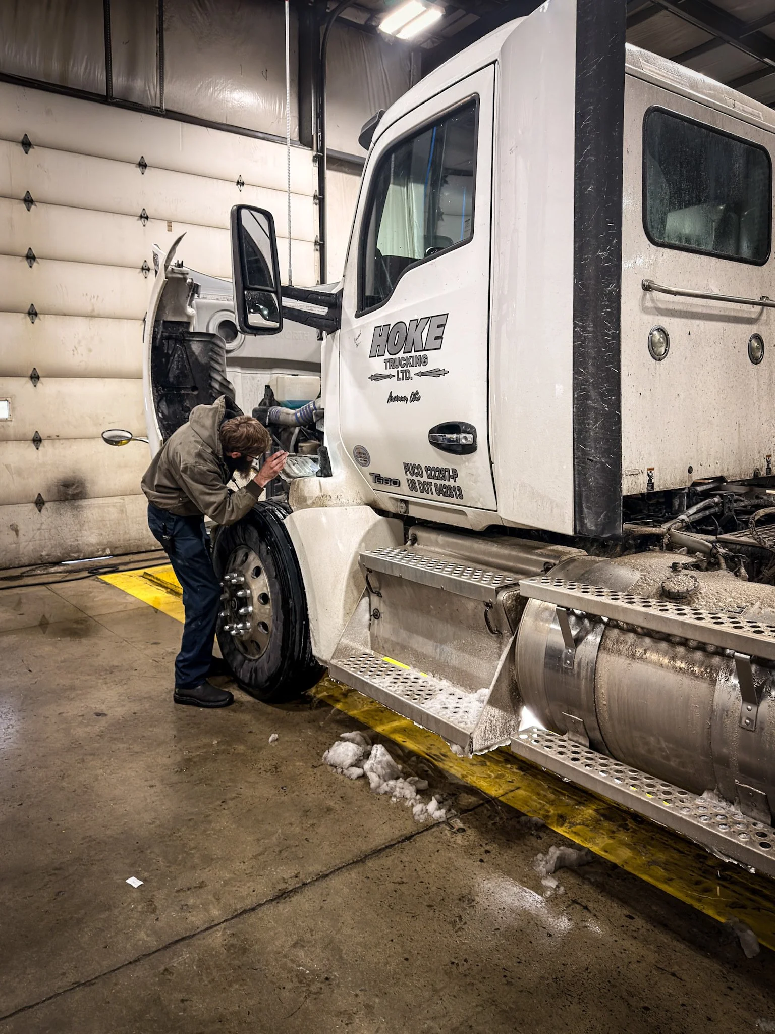 A mechanic inspecting the front wheel of a large white semi-truck inside a garage, with snow on the floor nearby.