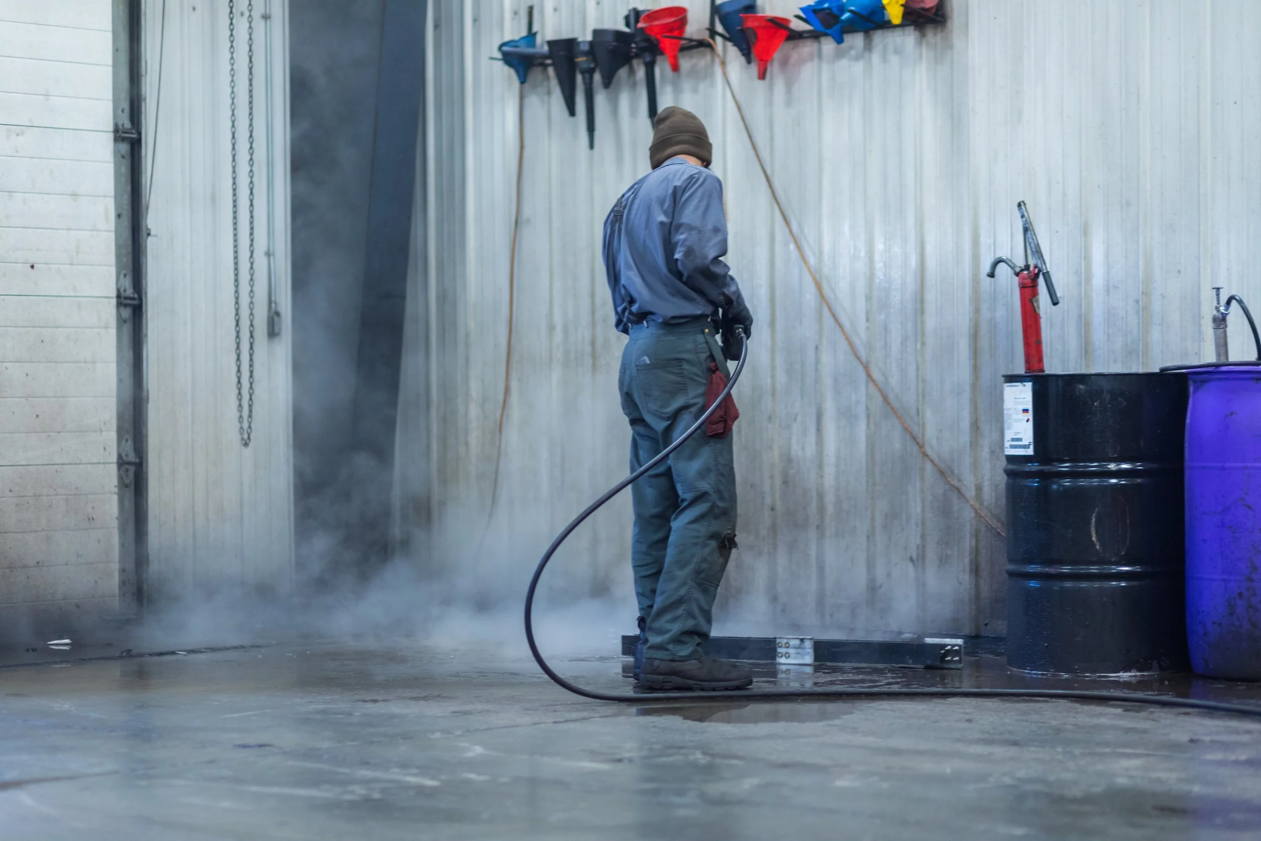 A worker in protective workwear pressure washing a concrete floor inside a garage or industrial space.
