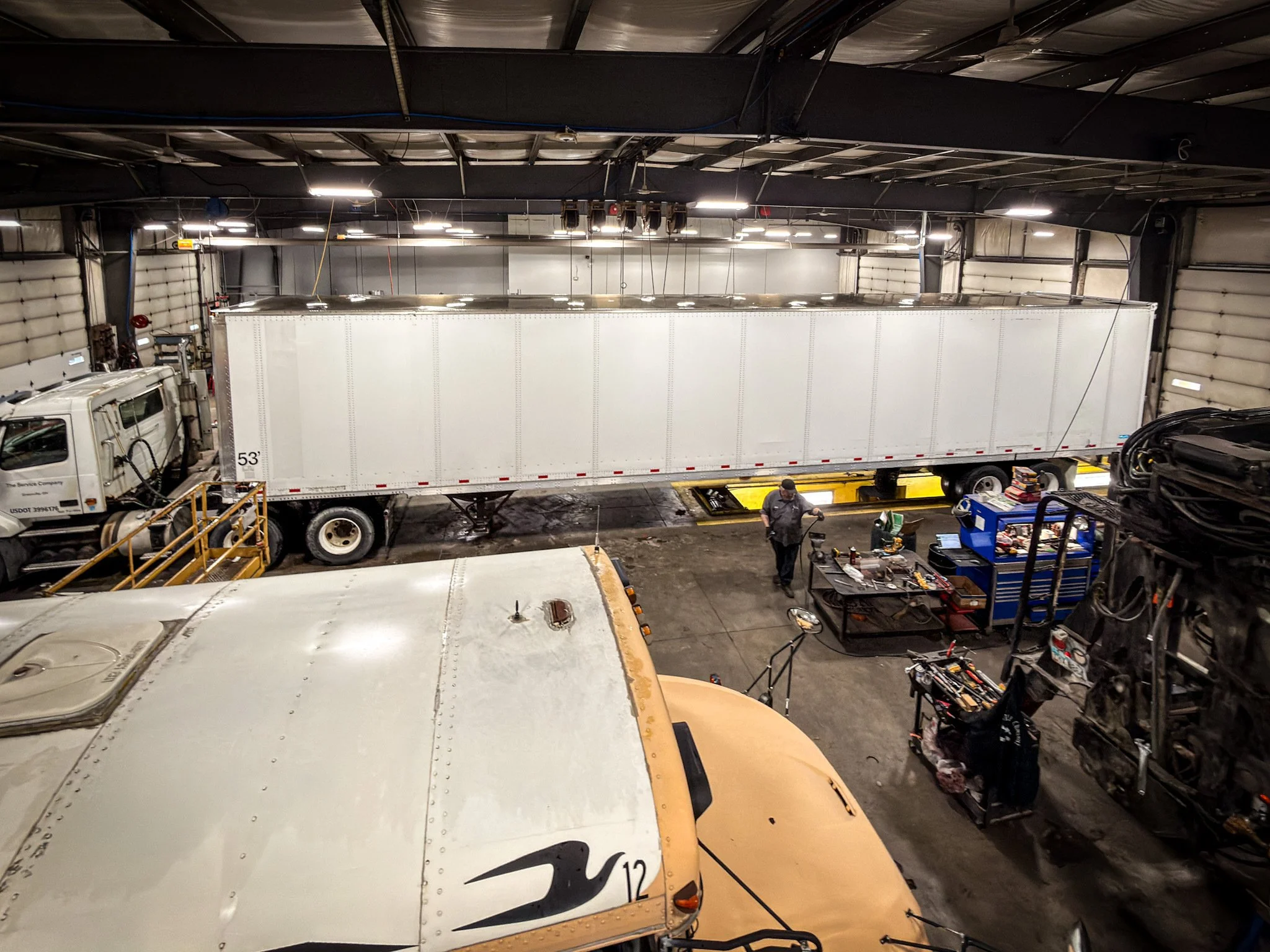 Inside a garage with trucks, including one with a white trailer, and tools and equipment on workbenches.