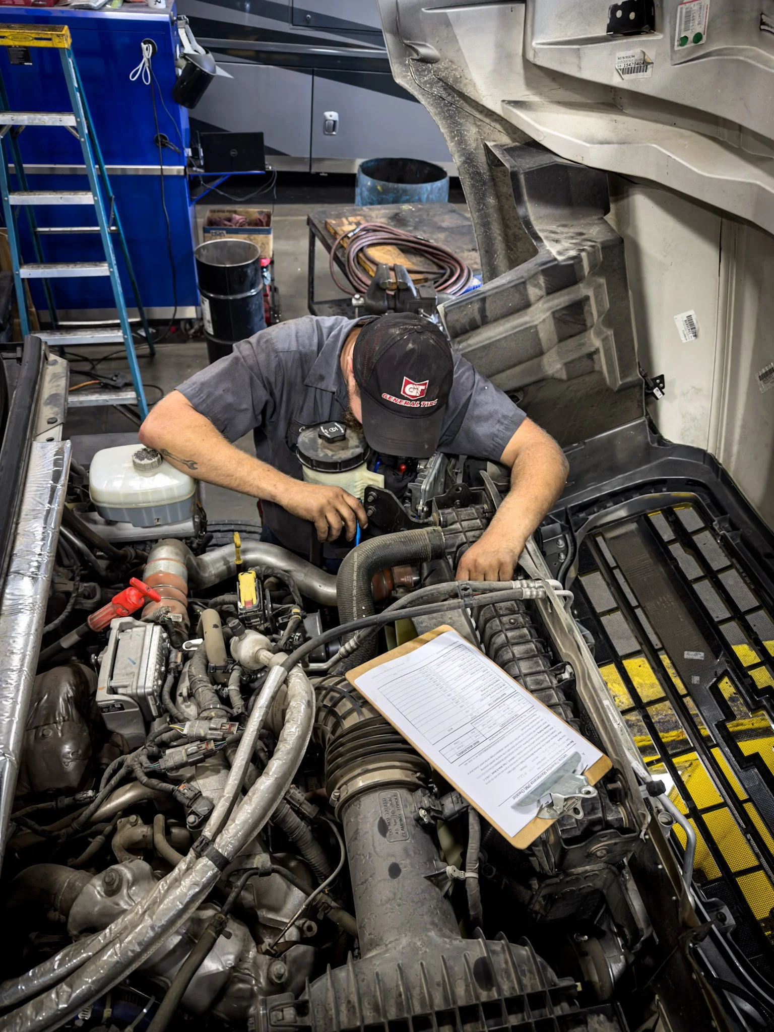 A mechanic working on a large engine inside a vehicle shop. The engine bay has various hoses, wires, and components. The mechanic is wearing a gray shirt and a black cap, leaning over the engine with tools in hand. There is a clipboard with papers on the engine.