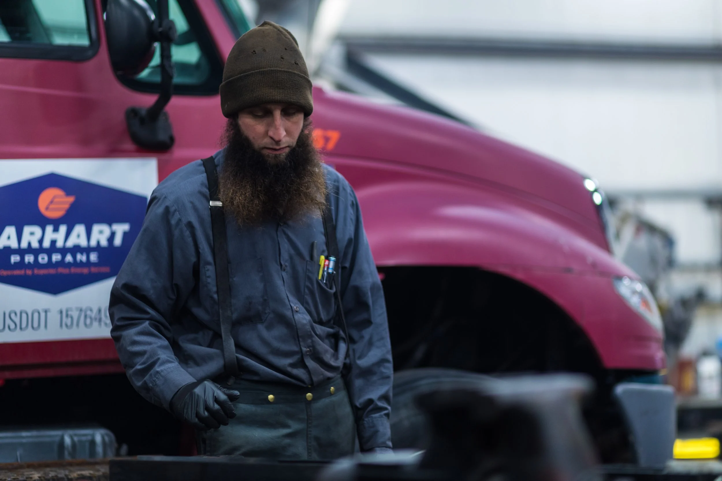 A man with a beard wearing a brown beanie, a blue shirt, and black gloves stands in front of a pink truck with a "Marhart Propane" logo on its side inside a garage or workshop.