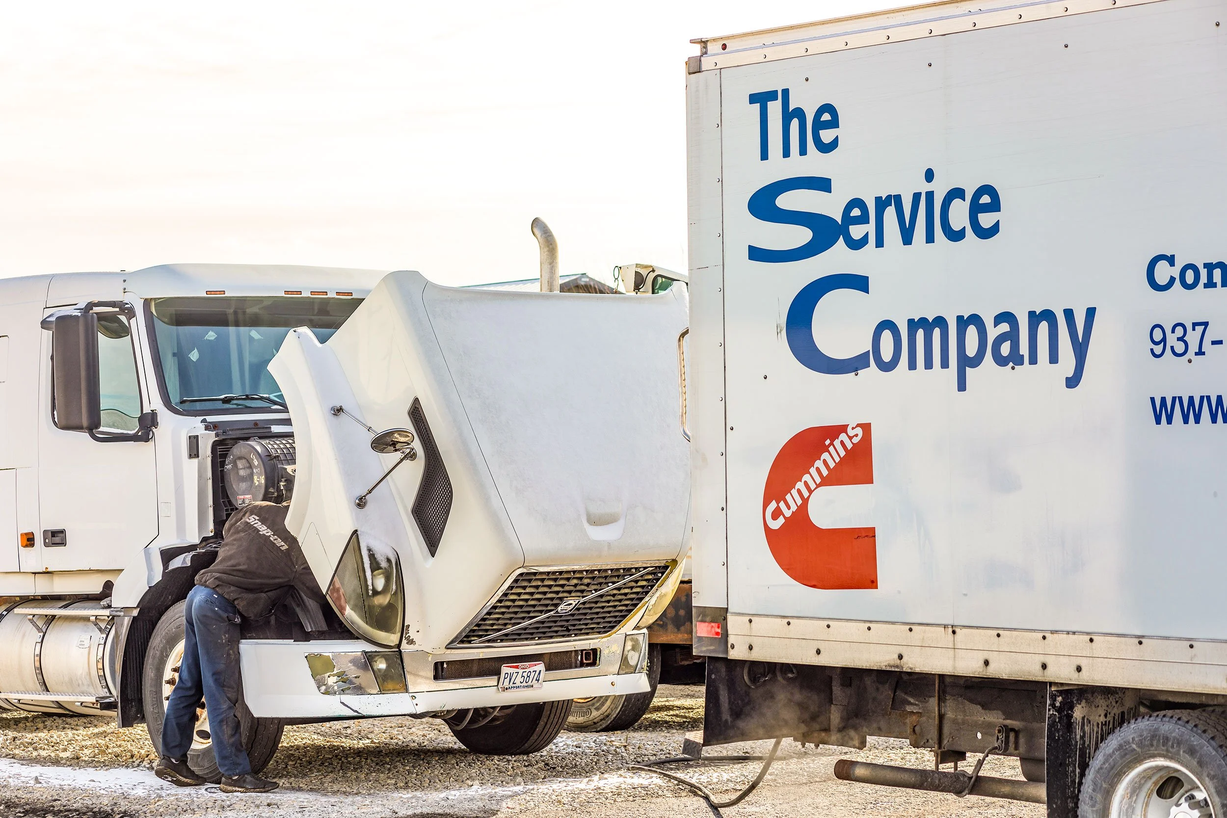 A mechanic inspecting a semi-truck's engine with a white box truck parked nearby, branded with 'The Service Company' and Cummins logo.