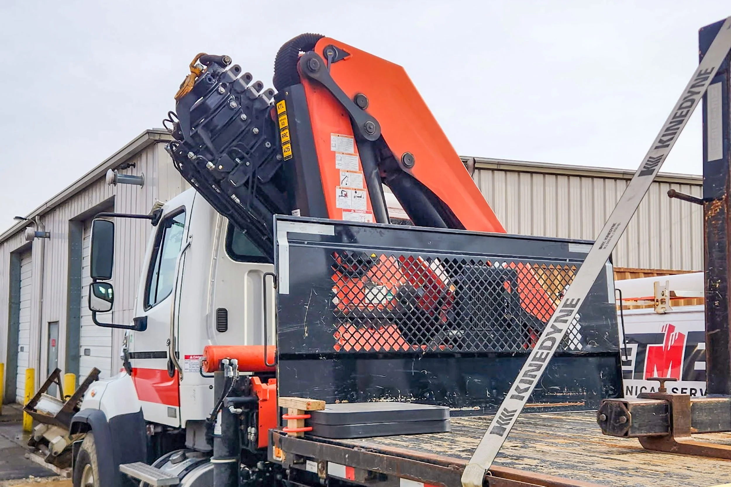 A white utility truck with an orange crane arm mounted on the back, parked outside a warehouse with multiple large doors.