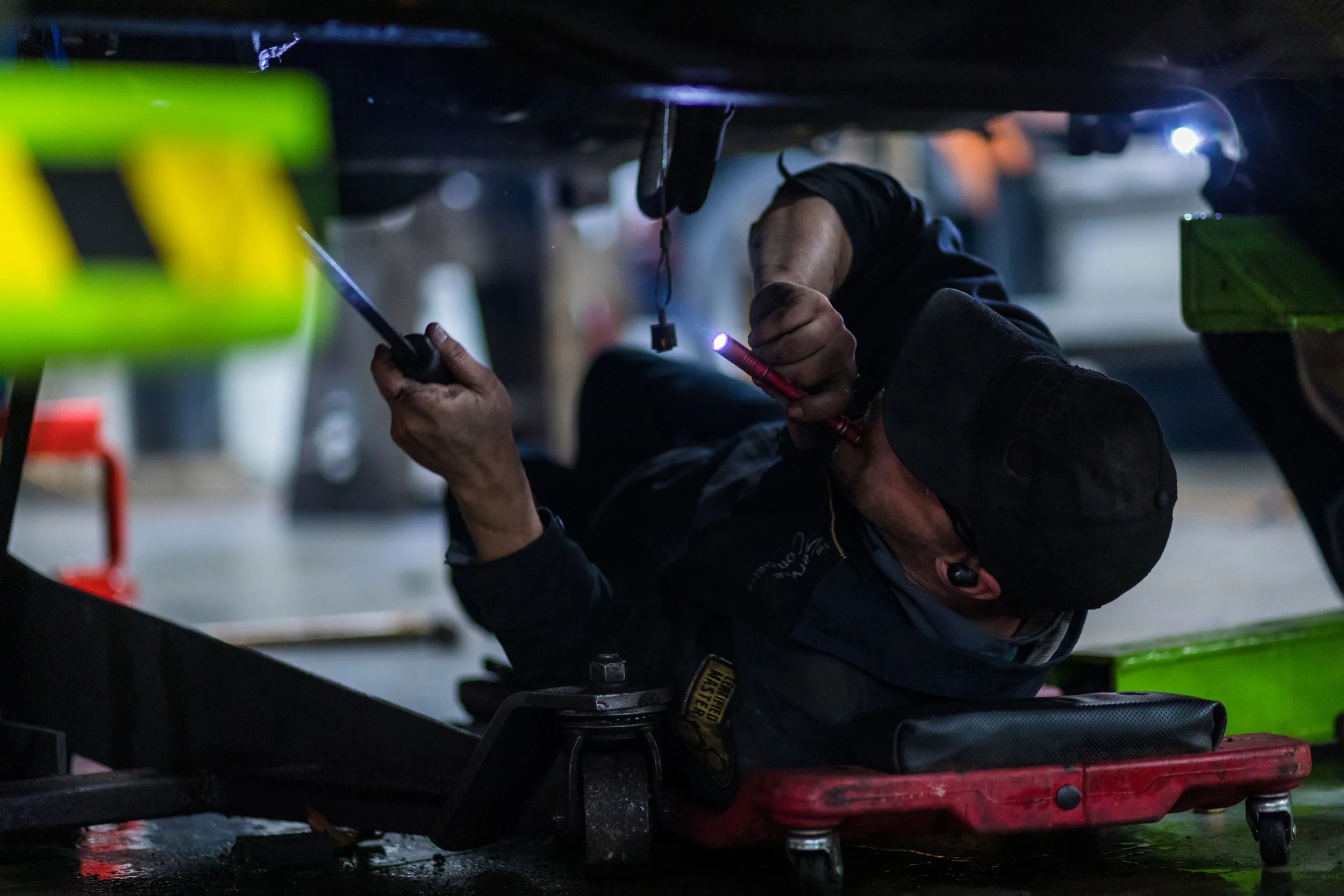 A mechanic working under a vehicle, lying on a creeper and using a flashlight to inspect the underside.