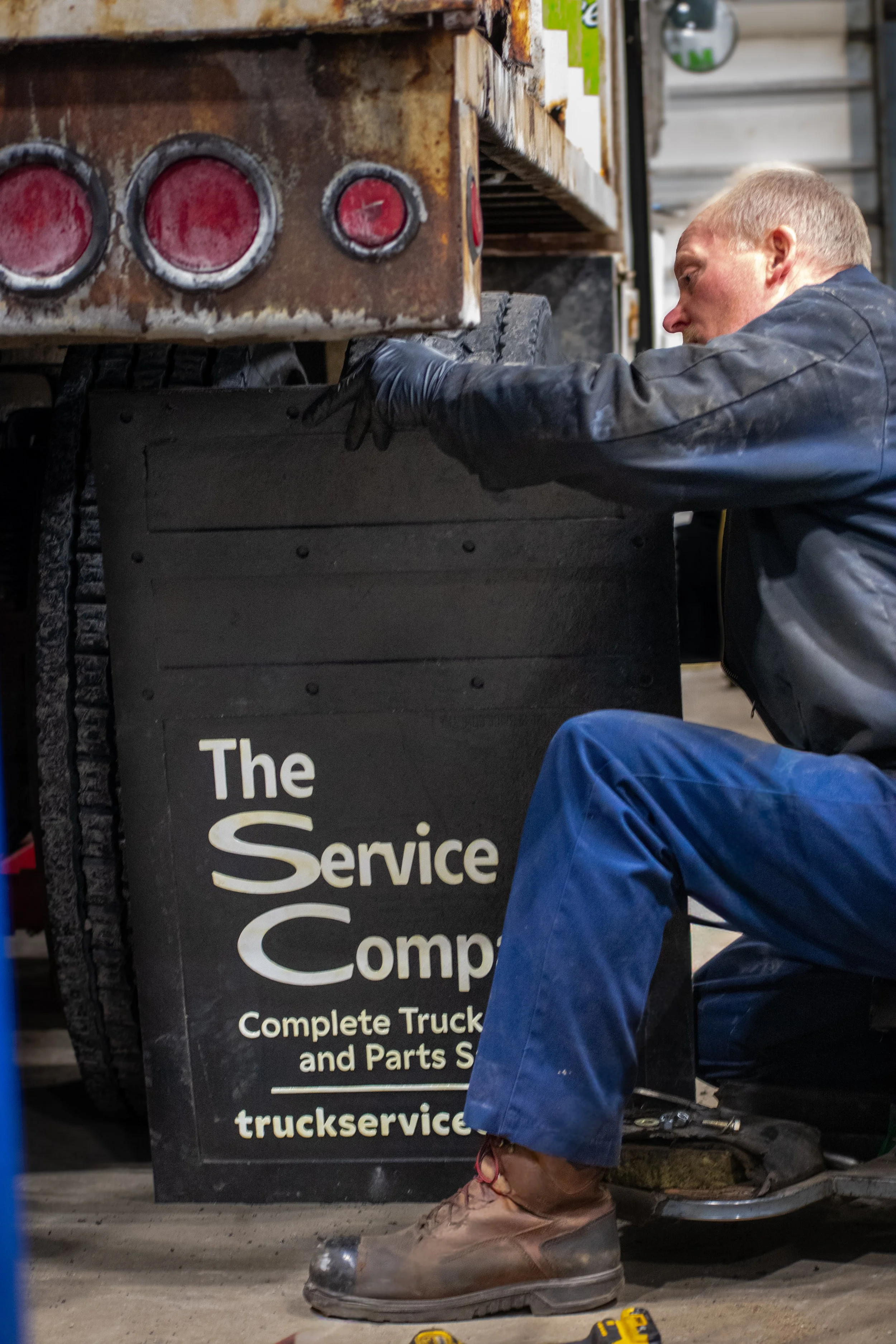 A man working underneath a truck, with a workshop sign that reads 'The Service Company' and 'Complete Truck and Parts Service'.