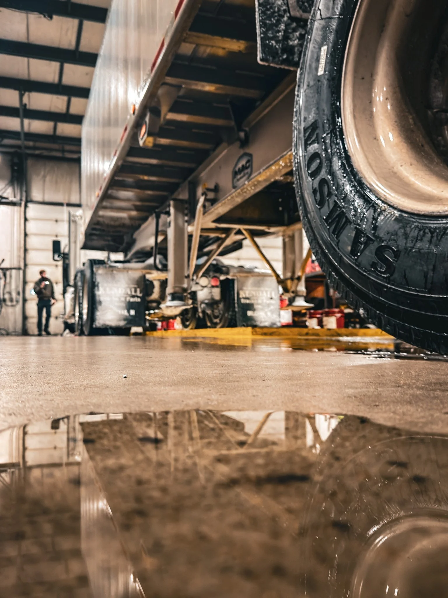View of the inside of an aircraft maintenance hangar seen from the ground, showing a large aircraft lifted on a lift and a tire with the word 'SHISO' on it in the foreground, with maintenance personnel and equipment in the background.