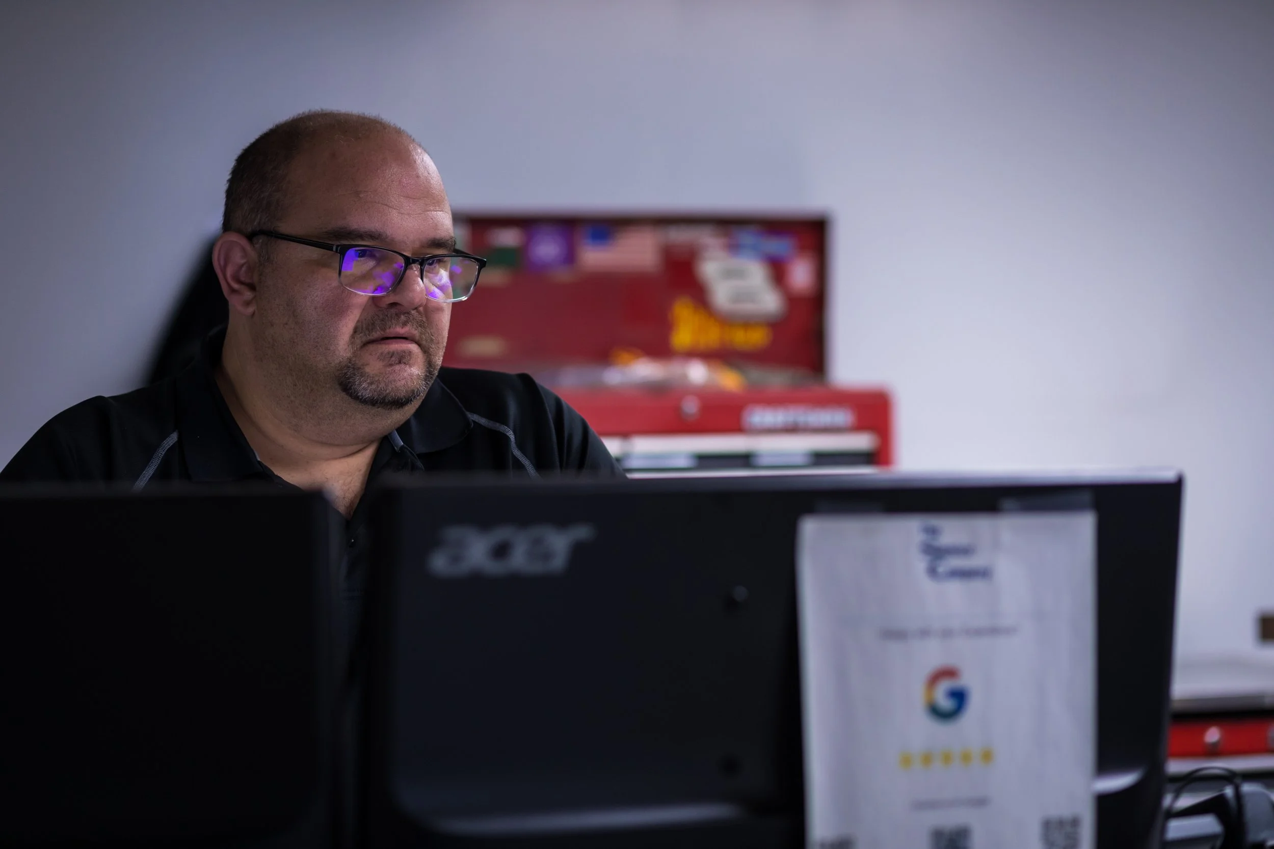 Man with glasses working at a computer in an office setting, with a red toolbox in the background.