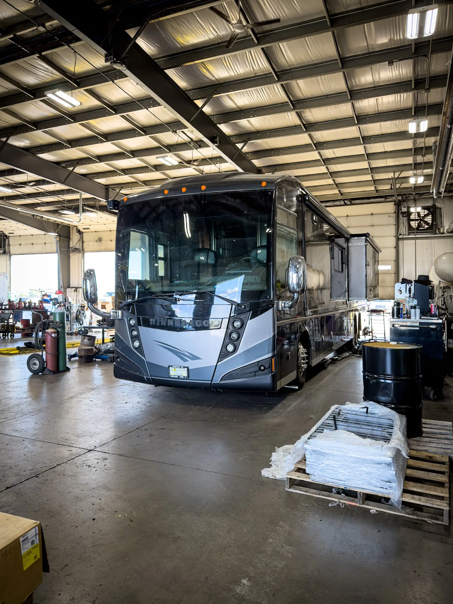 A large black and gray Winnebago motorhome inside a spacious industrial garage or workshop with tools, equipment, and supplies around.