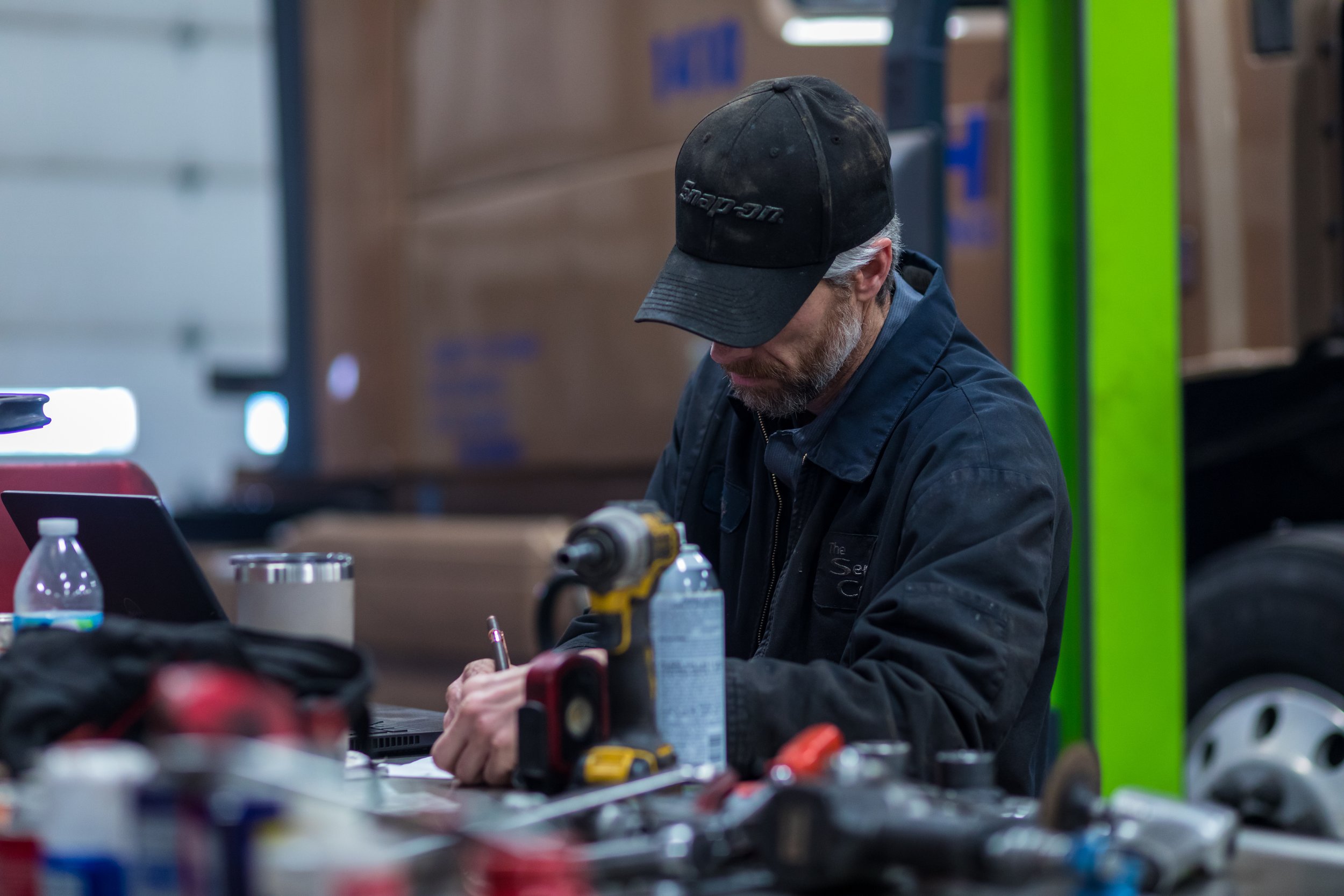 A man working at a cluttered workshop table, wearing a black cap and jacket, with tools and a laptop on the table.