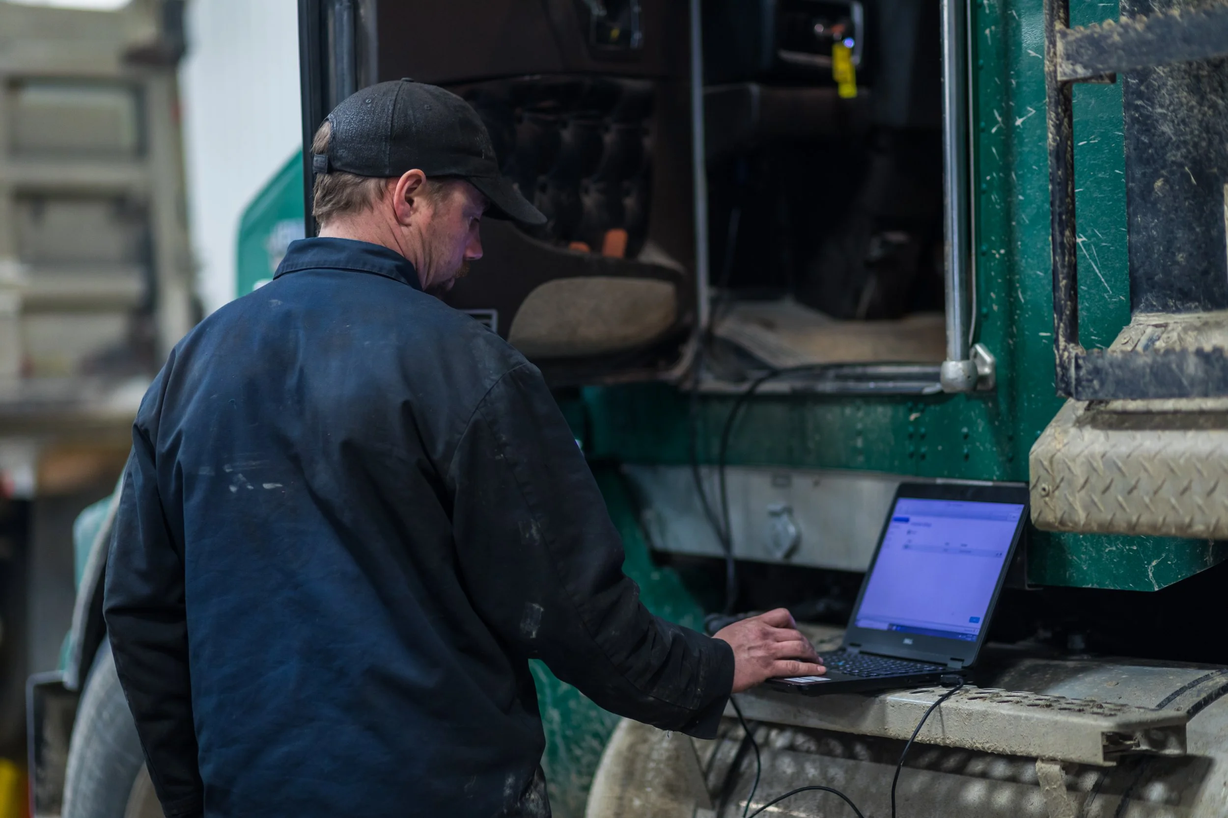 A man in a black jacket and black cap working on a laptop at a work site next to a large, green industrial vehicle.