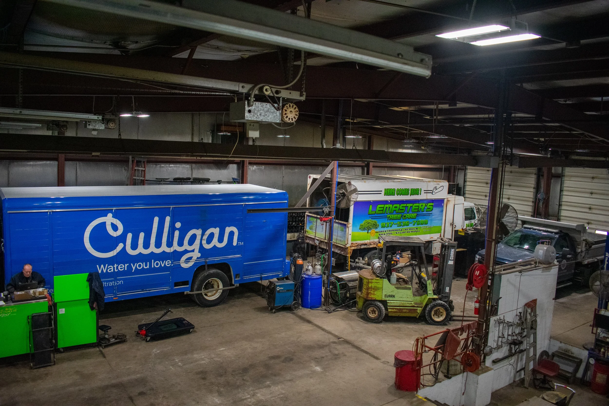 Inside a warehouse, several service trucks including Culligan water delivery truck and Lemaster's Tree Care truck, a forklift, and various equipment and tools are visible.