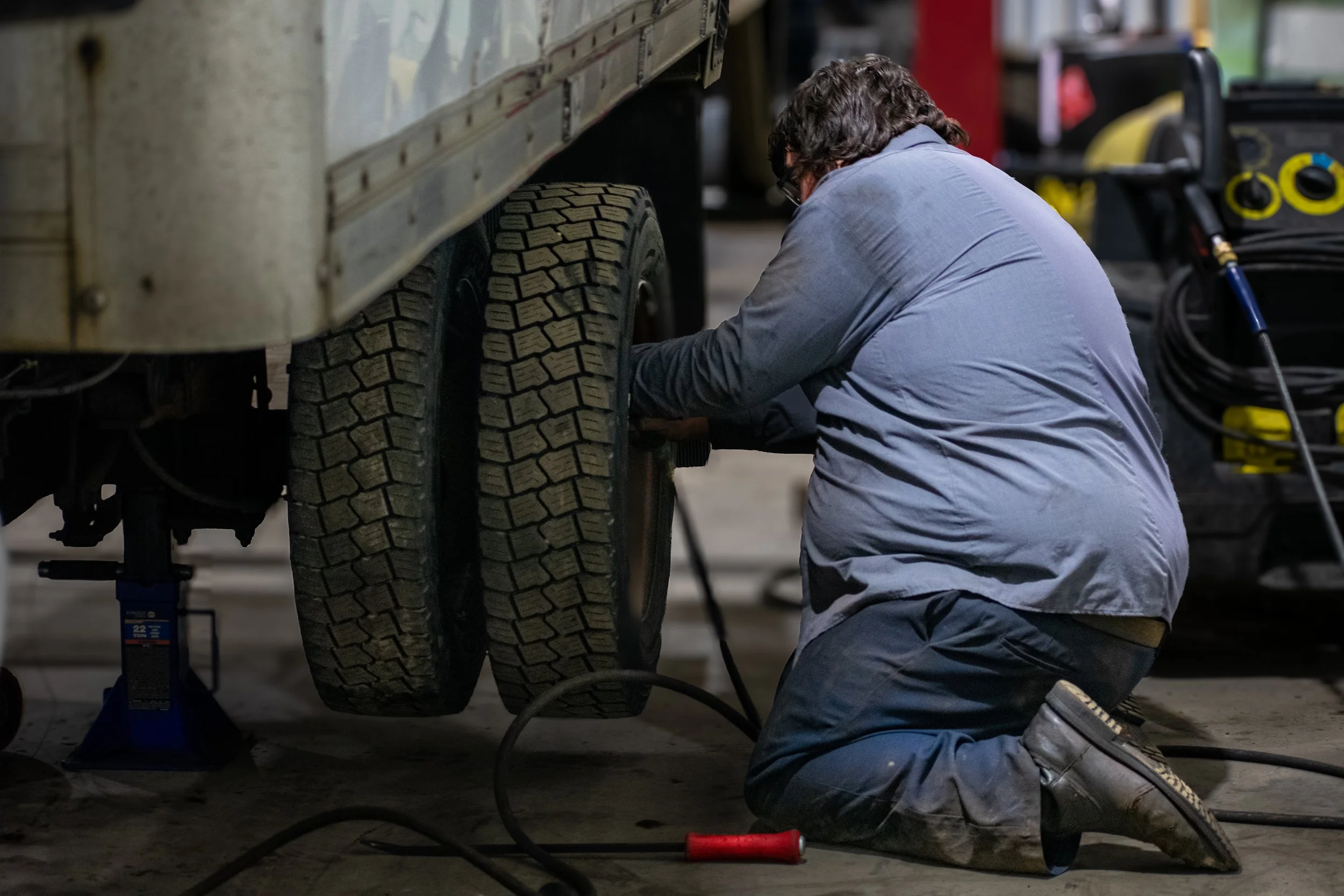A mechanic kneeling on the ground repairing two tires on a large vehicle in a garage or workshop environment.