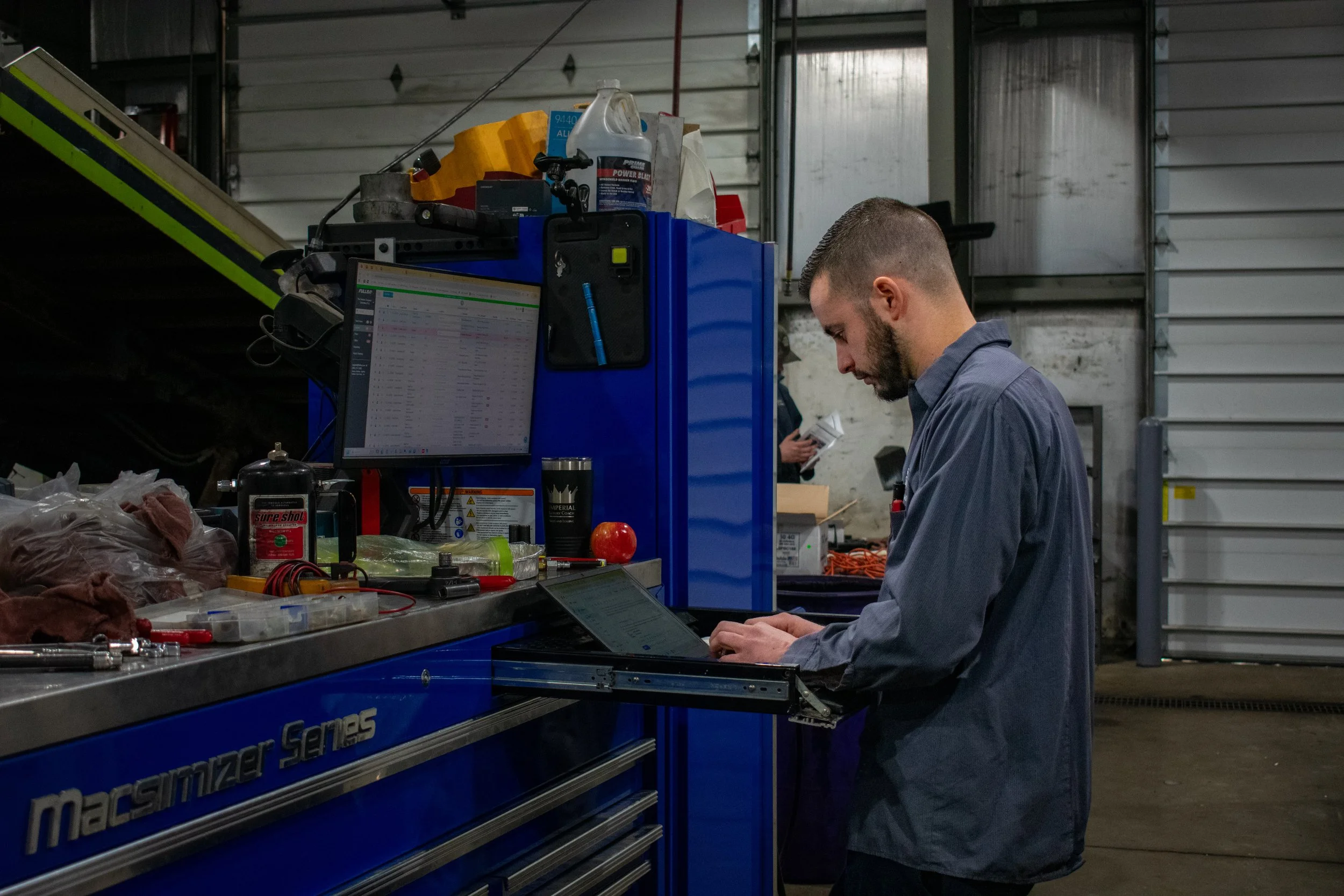 A man working at a blue Macstrizer Series tool chest in a garage or workshop, using a laptop with other tools and supplies on the work surface.