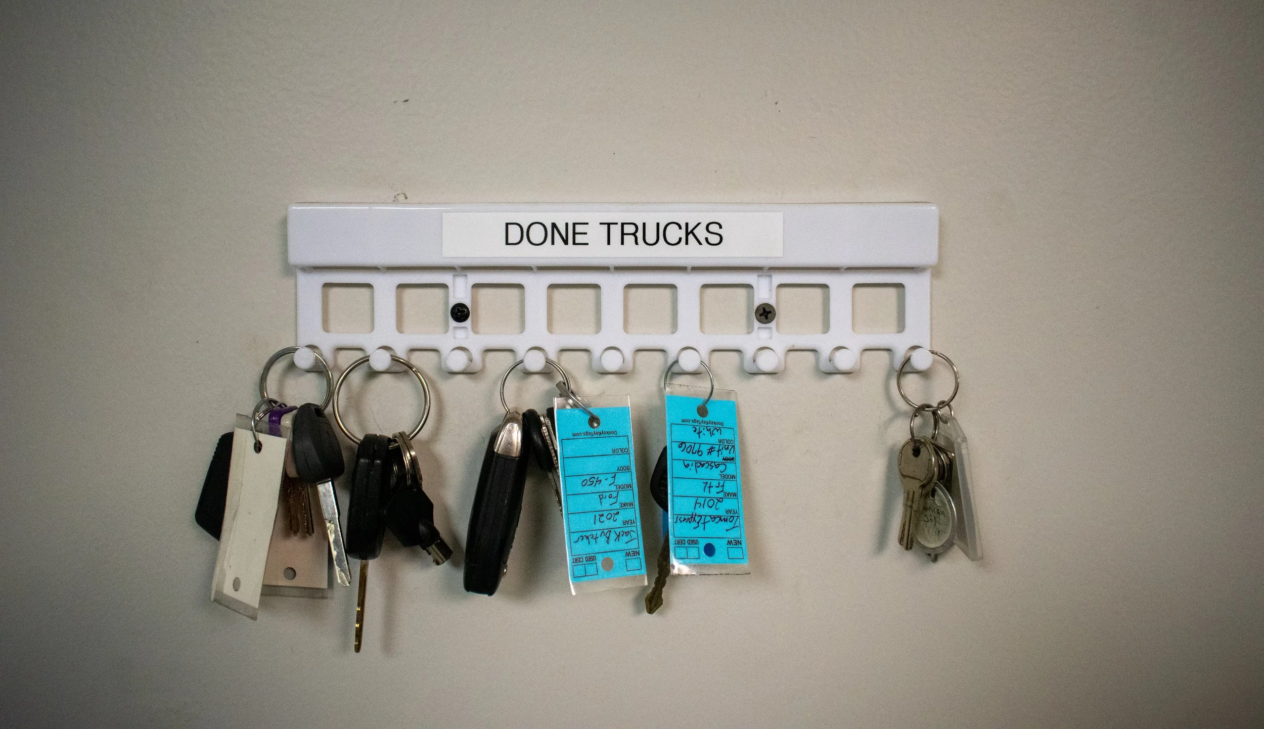 A white key rack labeled 'DONE TRUCKS' with several keys and key fobs hanging on it, mounted on a beige wall.