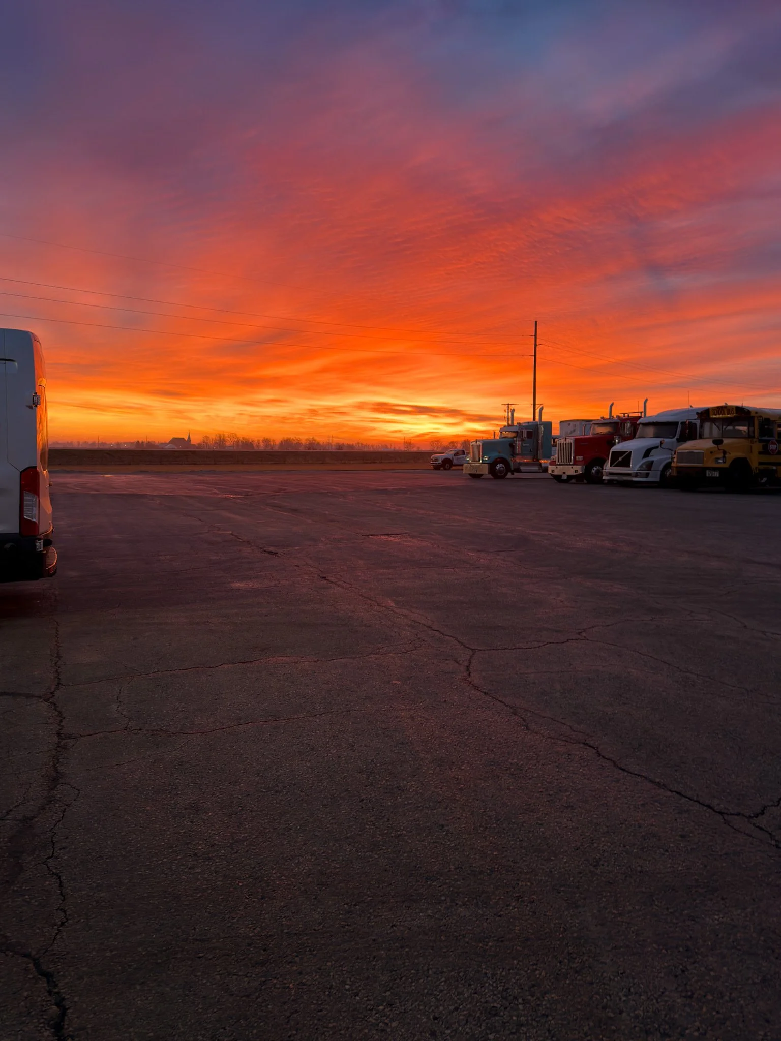 A parking lot during sunset with a colorful sky filled with orange, pink, and purple hues. Several trucks and vehicles are parked along the right side of the lot, with no people visible.