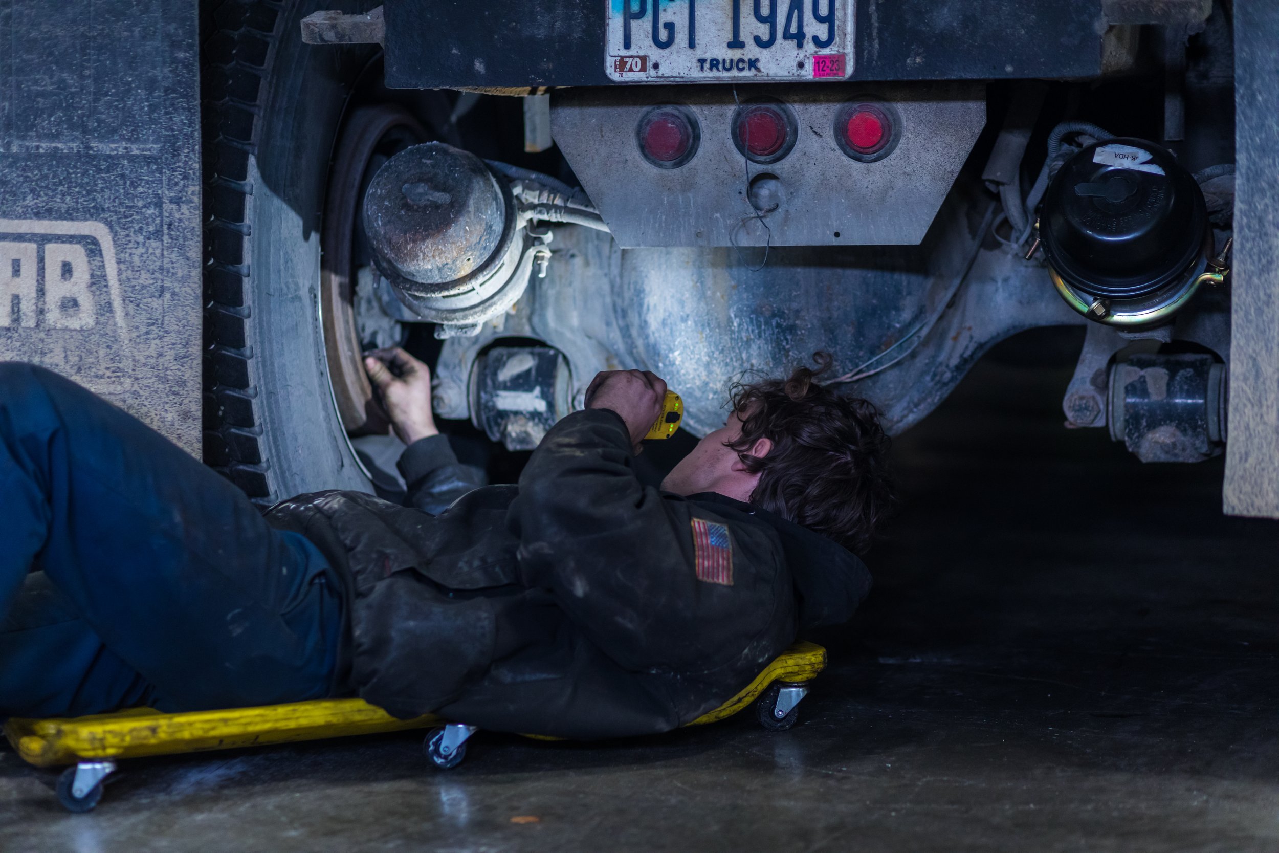 A person lying on a yellow creeper, working underneath a large, dusty industrial vehicle, possibly a truck or construction equipment, with tools and equipment around.