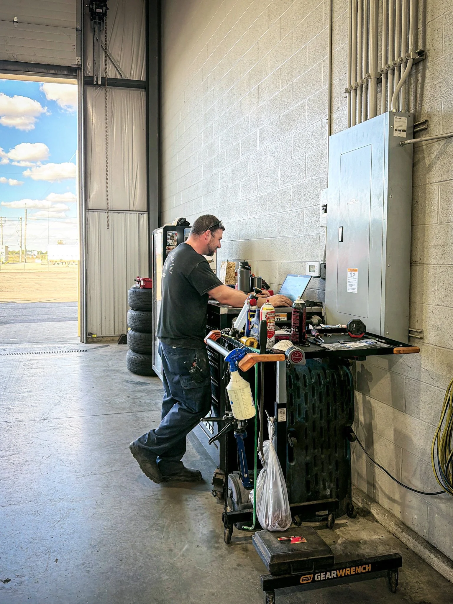 A man working at a workstation inside a garage or workshop, with tools, supplies, and tires nearby, and an open garage door showing the outdoors.