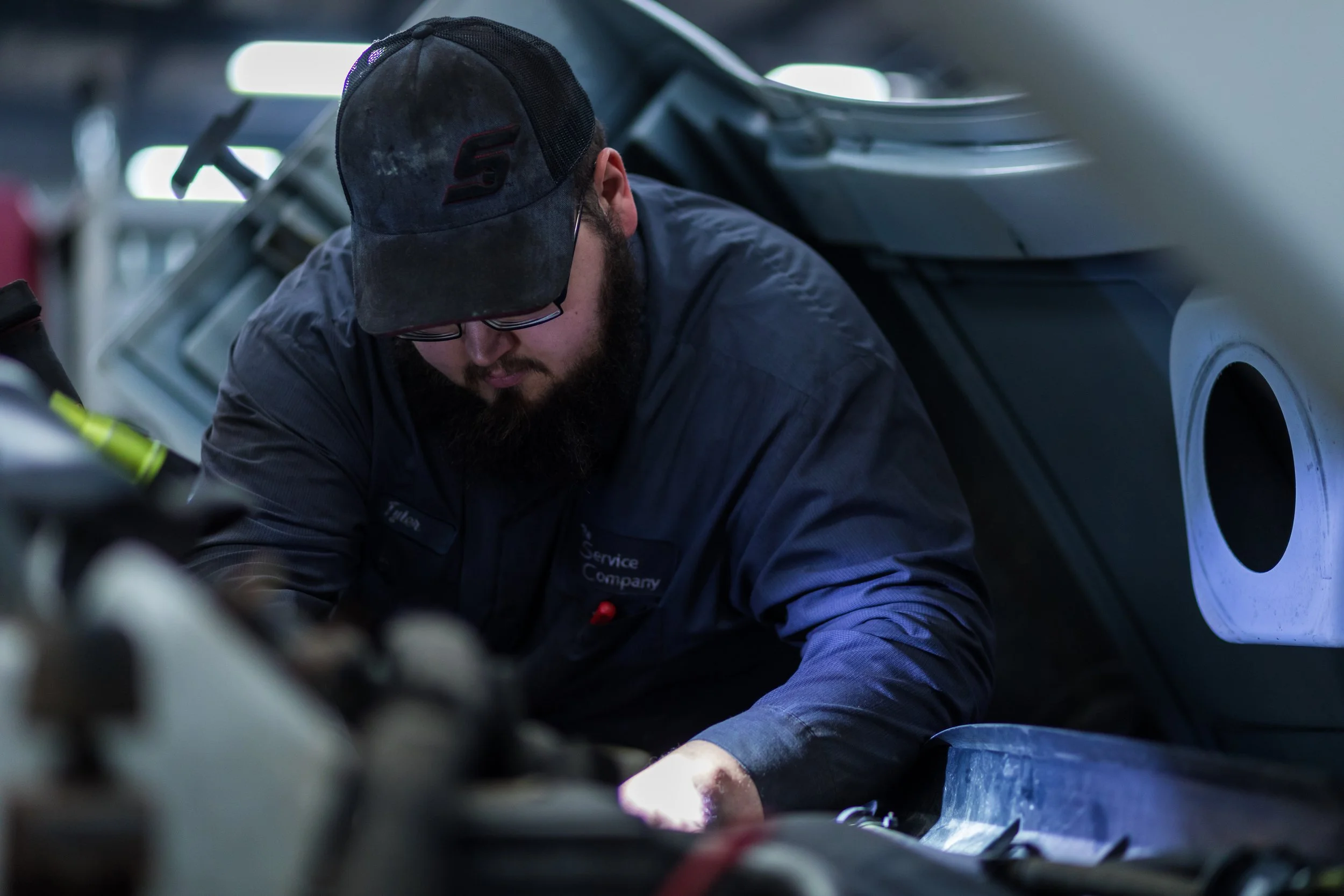 A man wearing glasses, a black cap, and a dark uniform working on a vehicle in an automotive repair shop.