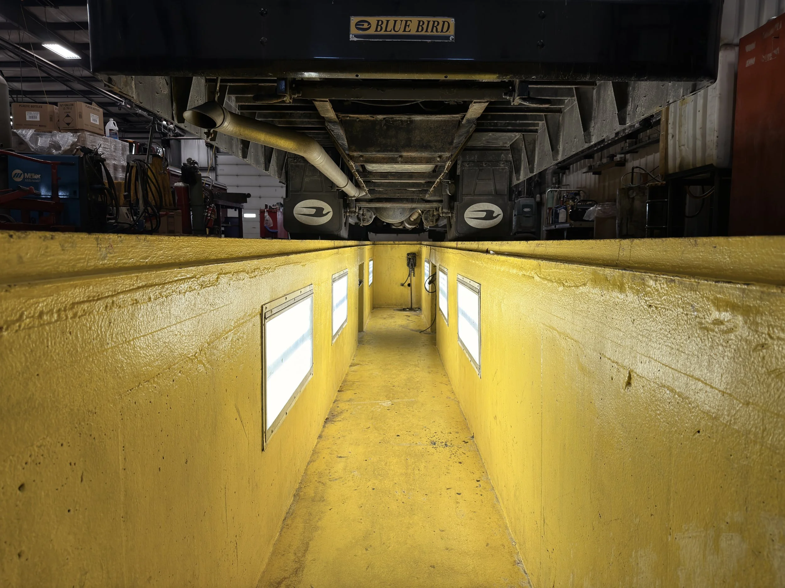 View from a workshop floor looking into an automotive lift with yellow guardrails and rectangular lights on the side, with workshop tools and equipment in the background.