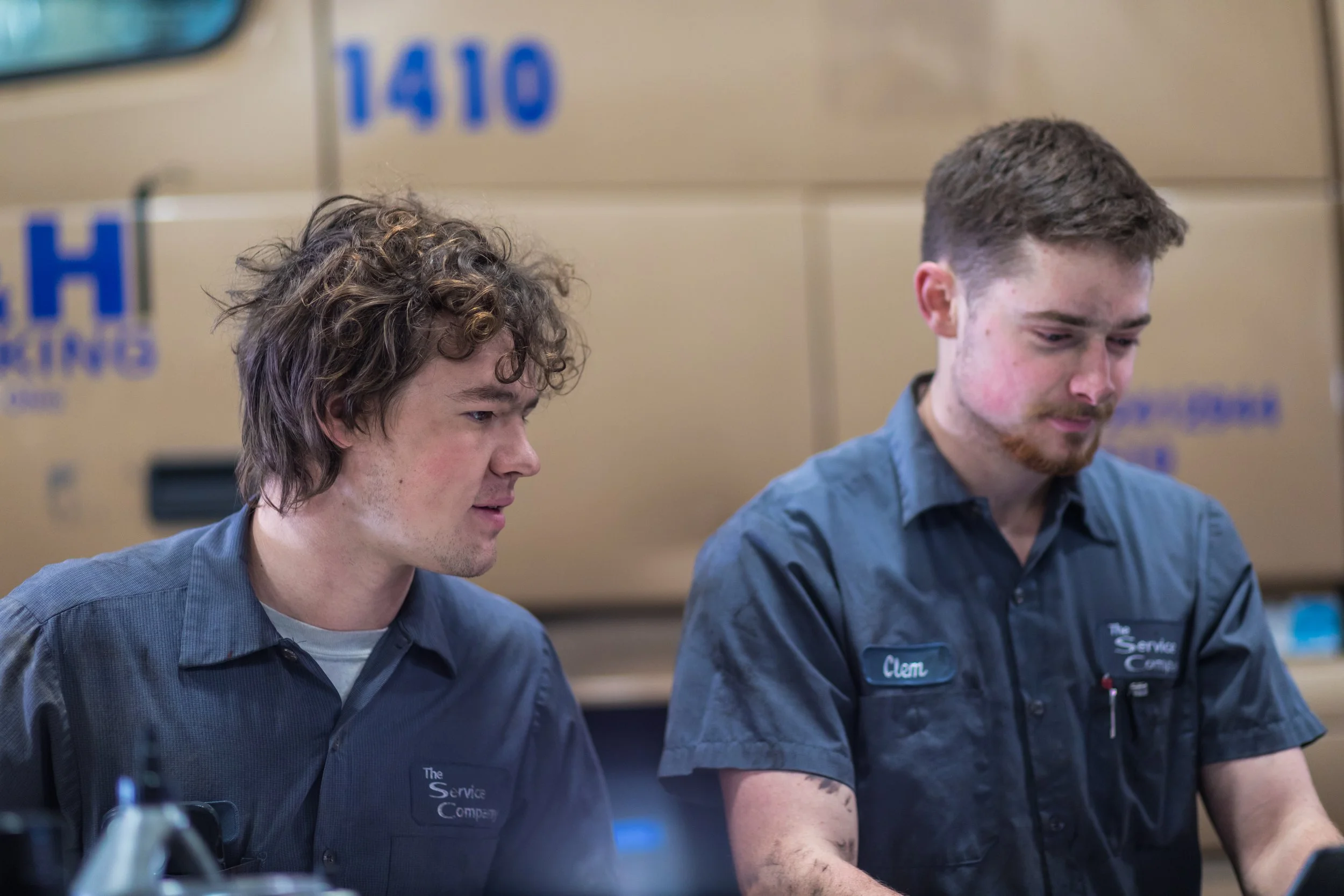 Two young men wearing blue work shirts sitting at a table, with a service company logo on their shirts, in an industrial setting with equipment and service trucks in the background.