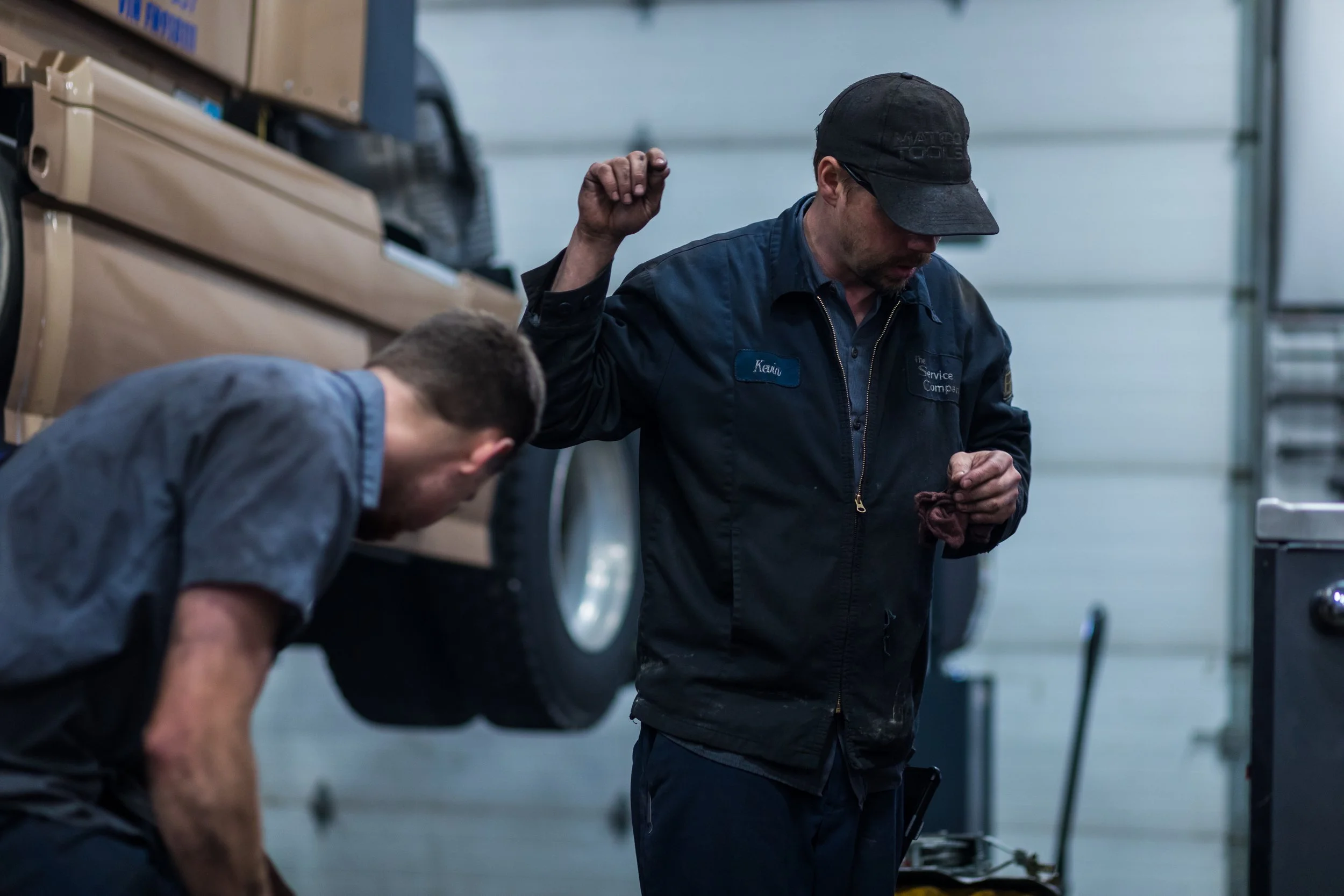 Two mechanics working in an automotive garage, one with a raised vehicle overhead and the other bending down.
