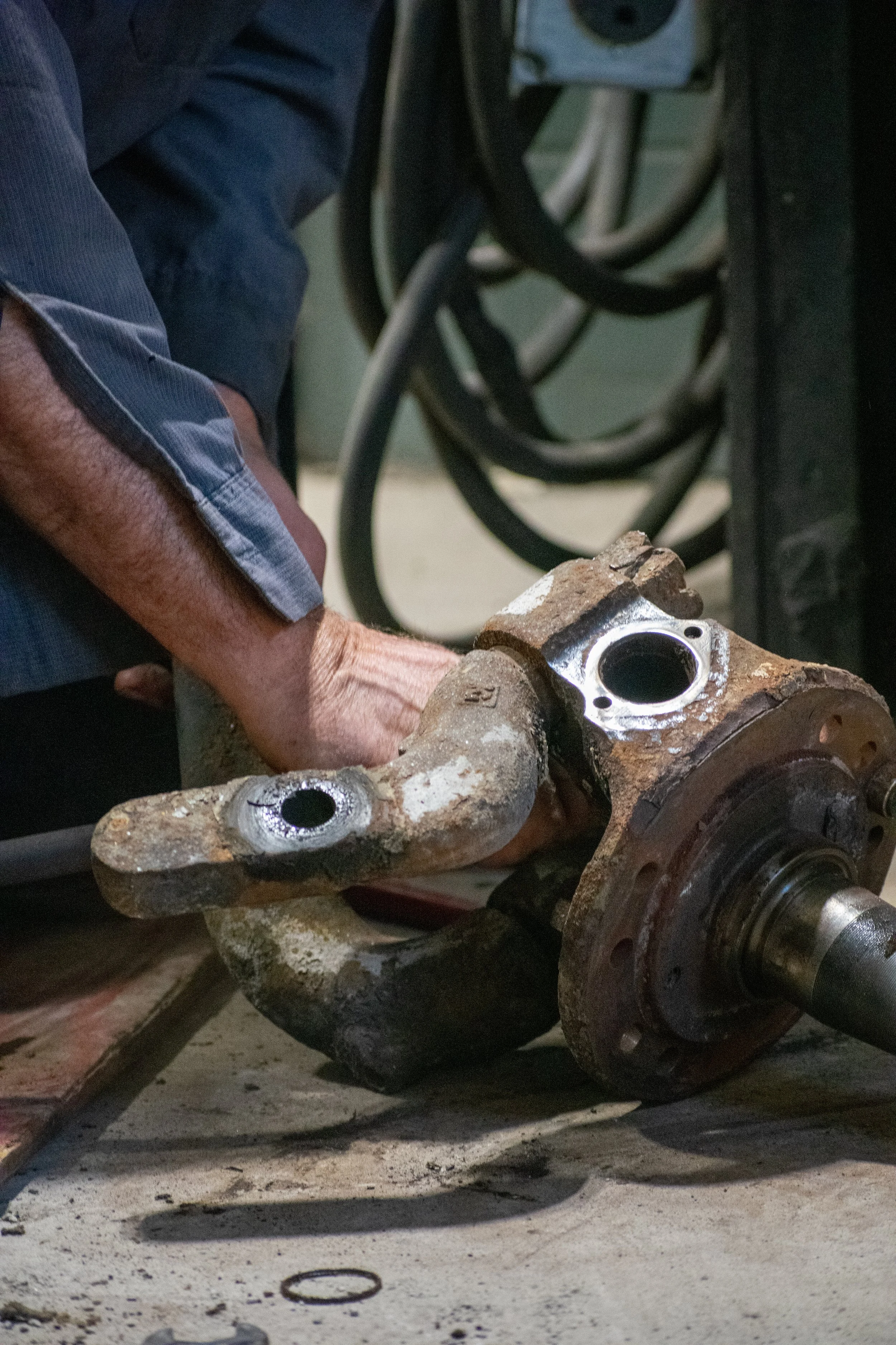 Close-up of a rusty, disassembled car part on a workbench, with a mechanic's arm in a blue shirt working on it.
