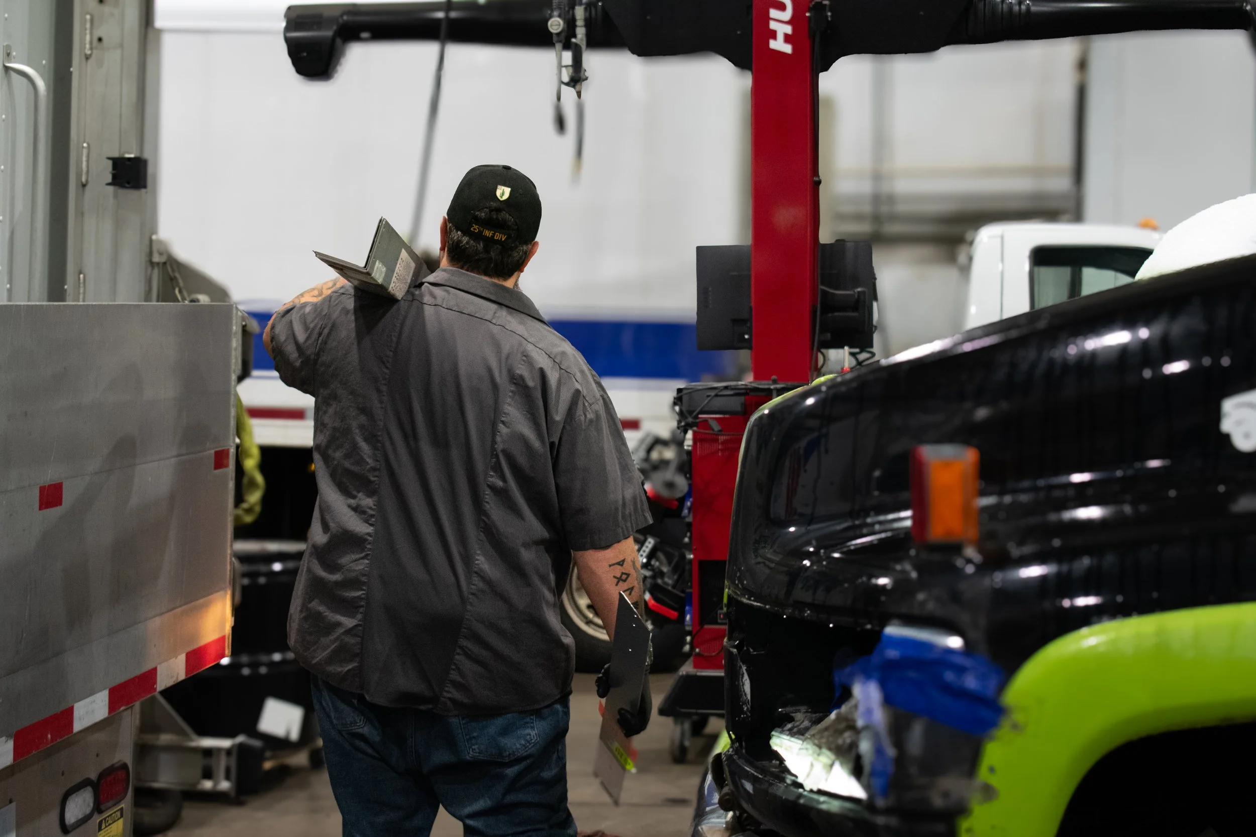 A man wearing a black hat and gray shirt working on vehicles inside a garage, with a black and yellow vehicle visible in the foreground and a red vehicle lift.