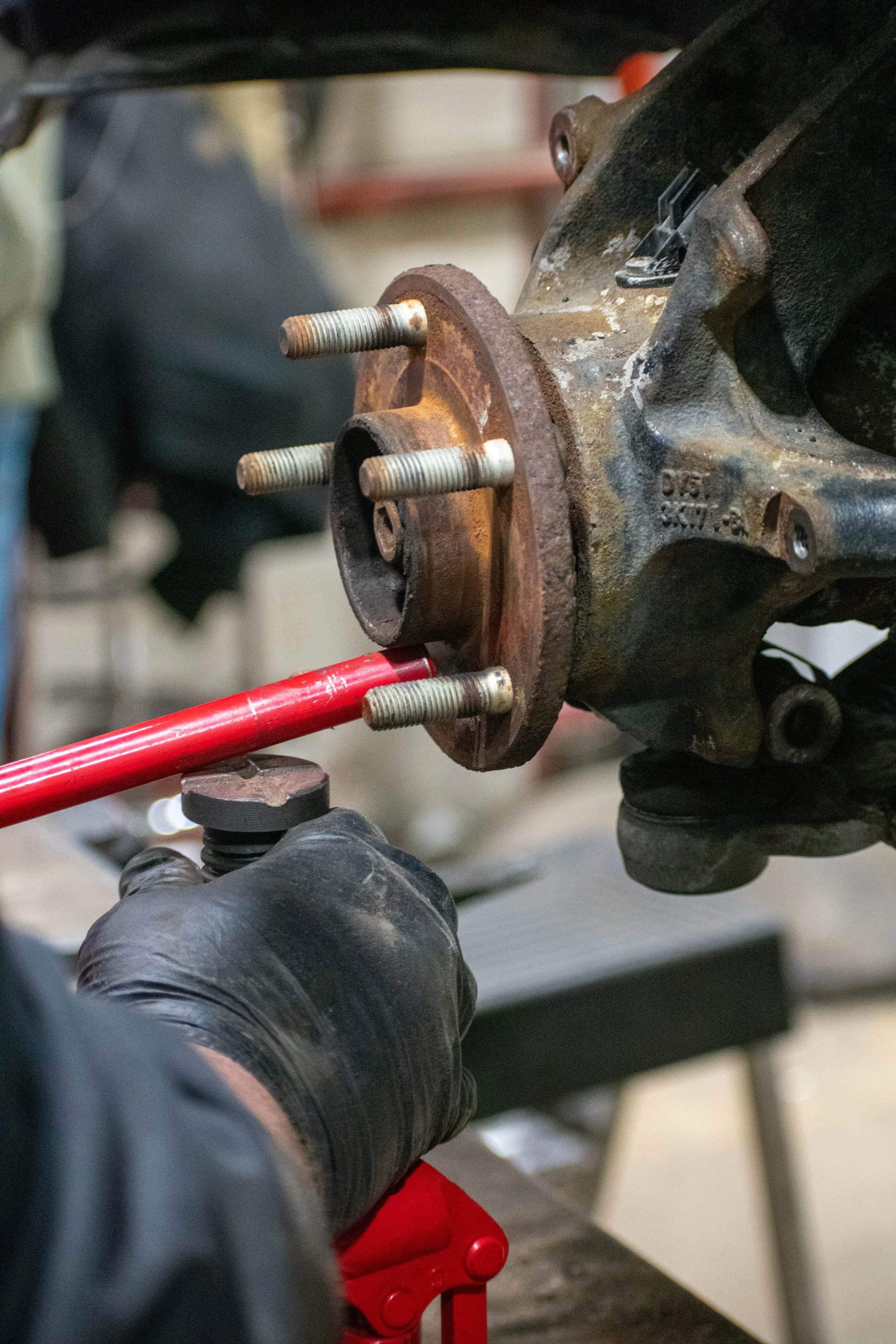A person wearing black gloves is working on a rusty car wheel hub, using a red tool for automotive repair.