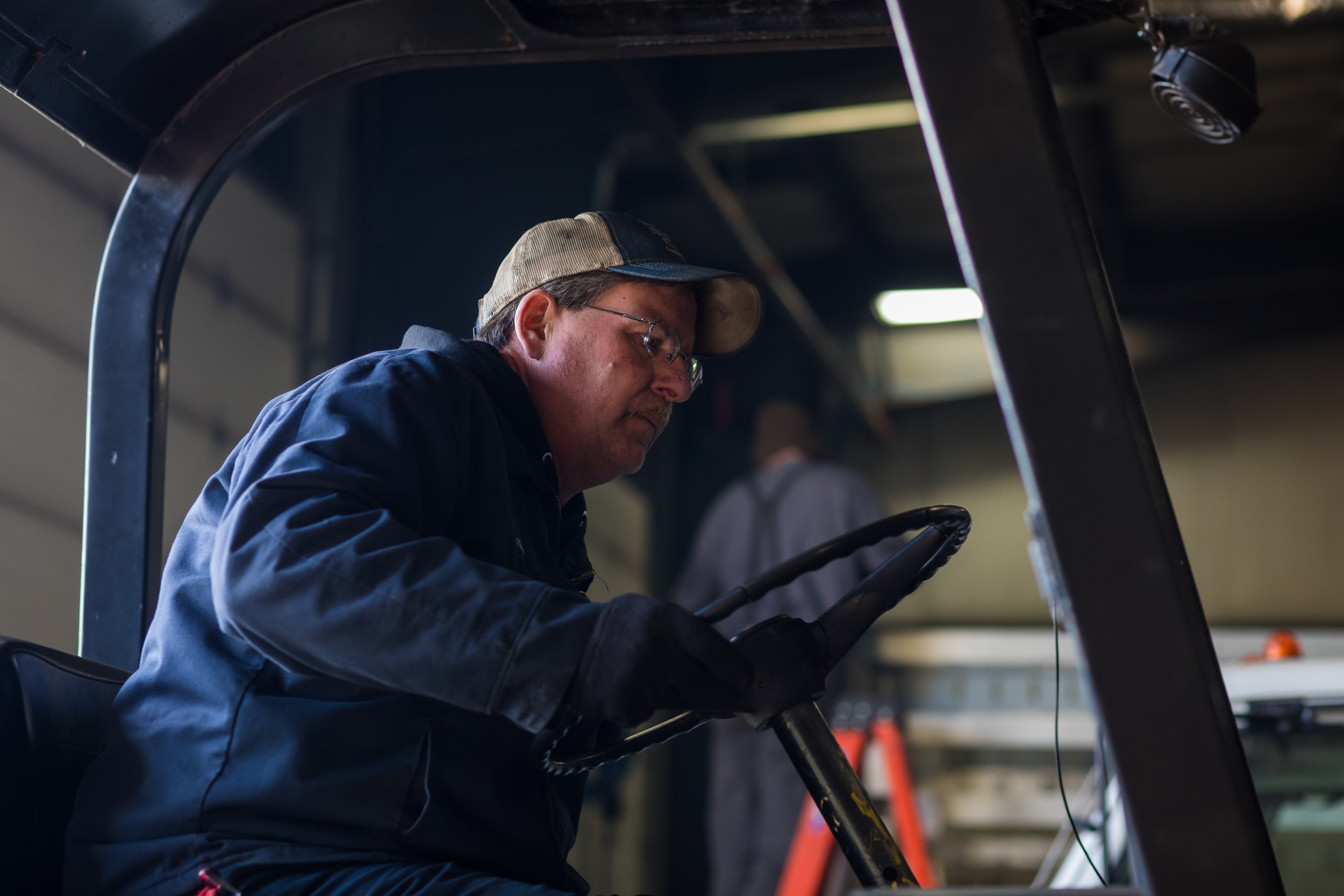 A man wearing a baseball cap, glasses, a dark jacket, and gloves is operating a forklift inside a warehouse.