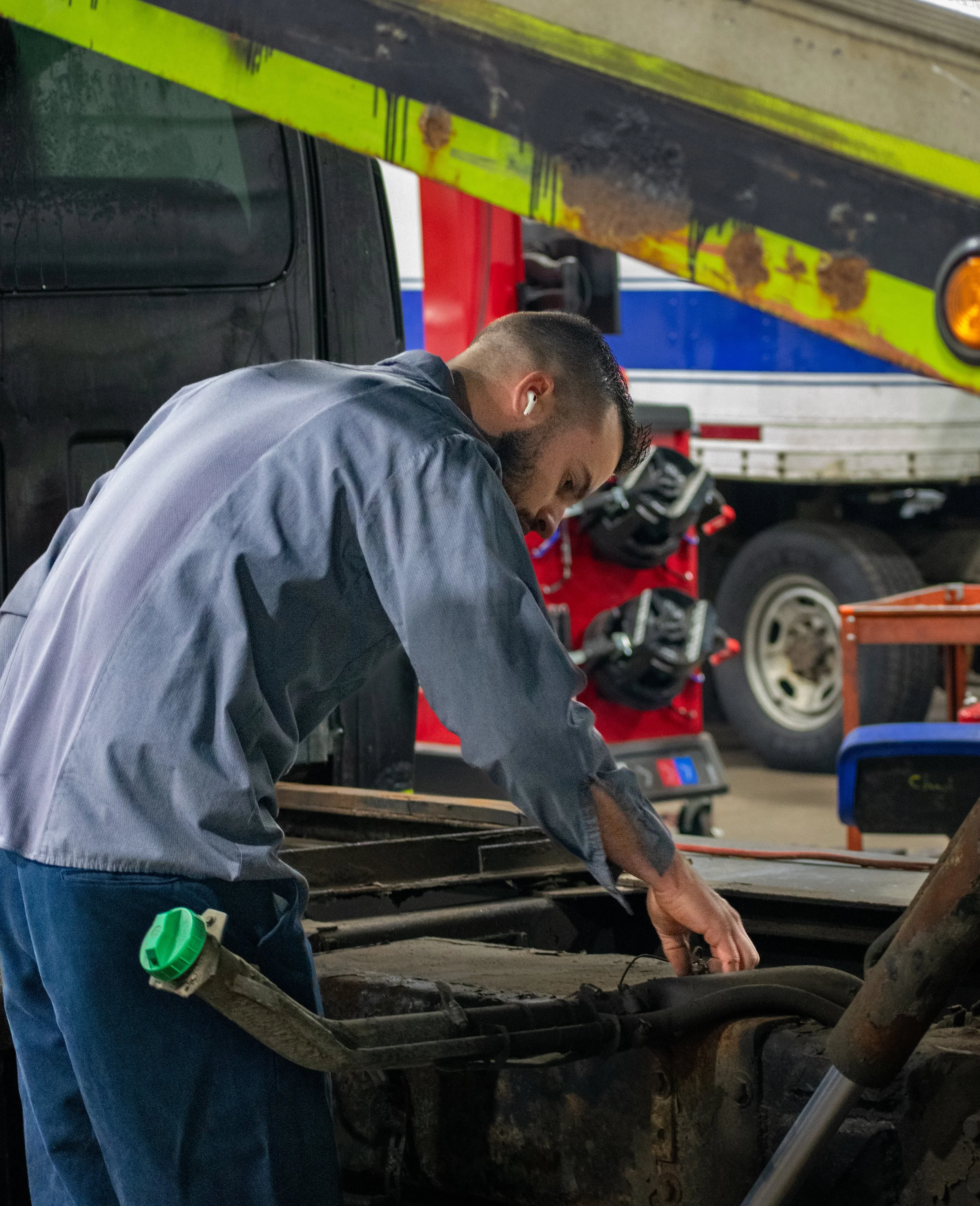 A man working on a vehicle engine in a garage, wearing a gray coverall, with tools and equipment around him.